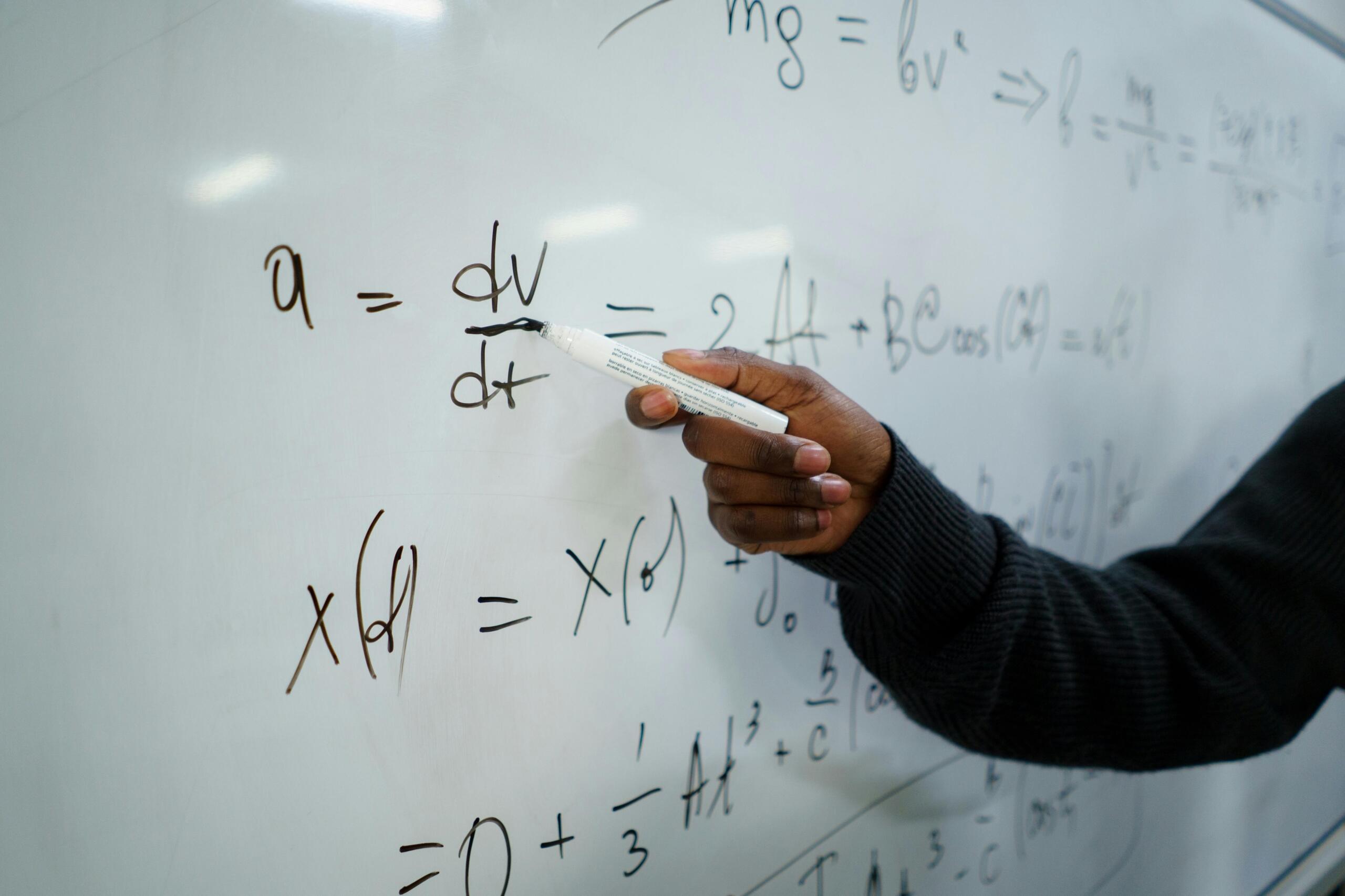 Mano de profesor escribiendo ecuaciones de cálculo diferencial y derivadas en pizarra blanca durante clase de matemáticas o física aplicada.
