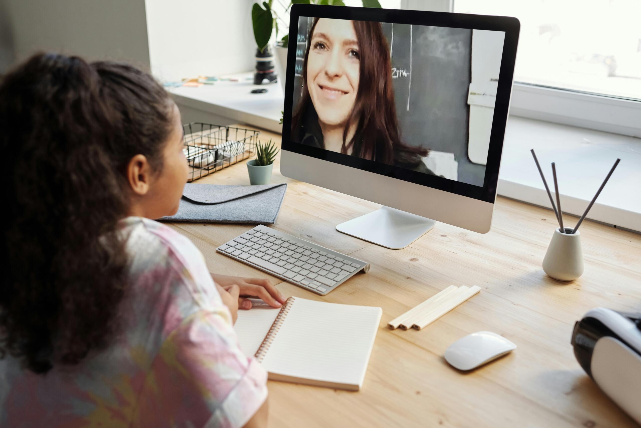 Estudiante adolescente recibiendo clase virtual frente a computadora de escritorio mientras observa a su profesora explicar lección desde aula con pizarra.
