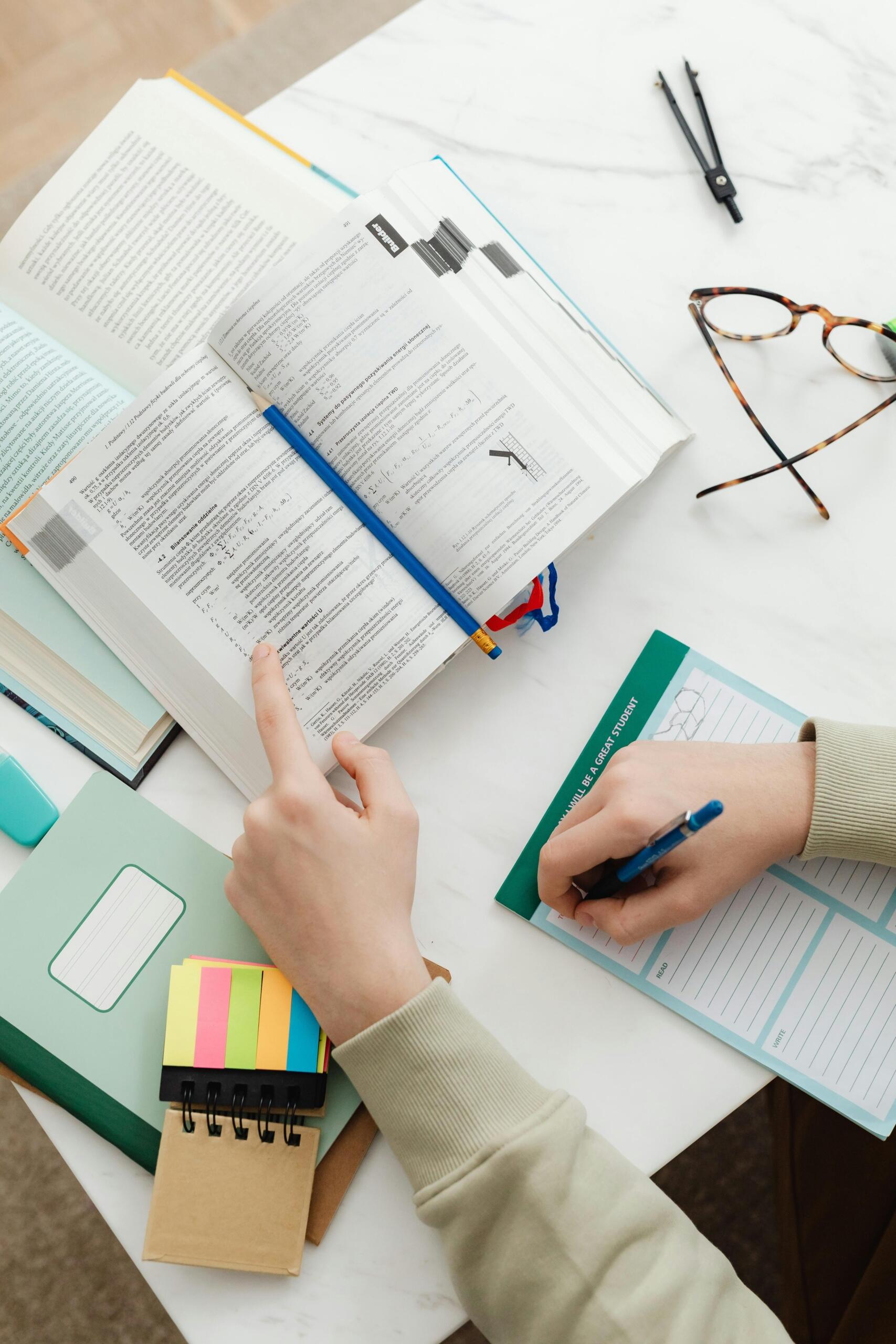 Vista cenital de estudiante consultando libro de texto técnico mientras toma apuntes en cuaderno, con notas adhesivas de colores, lentes y compás sobre escritorio de mármol.