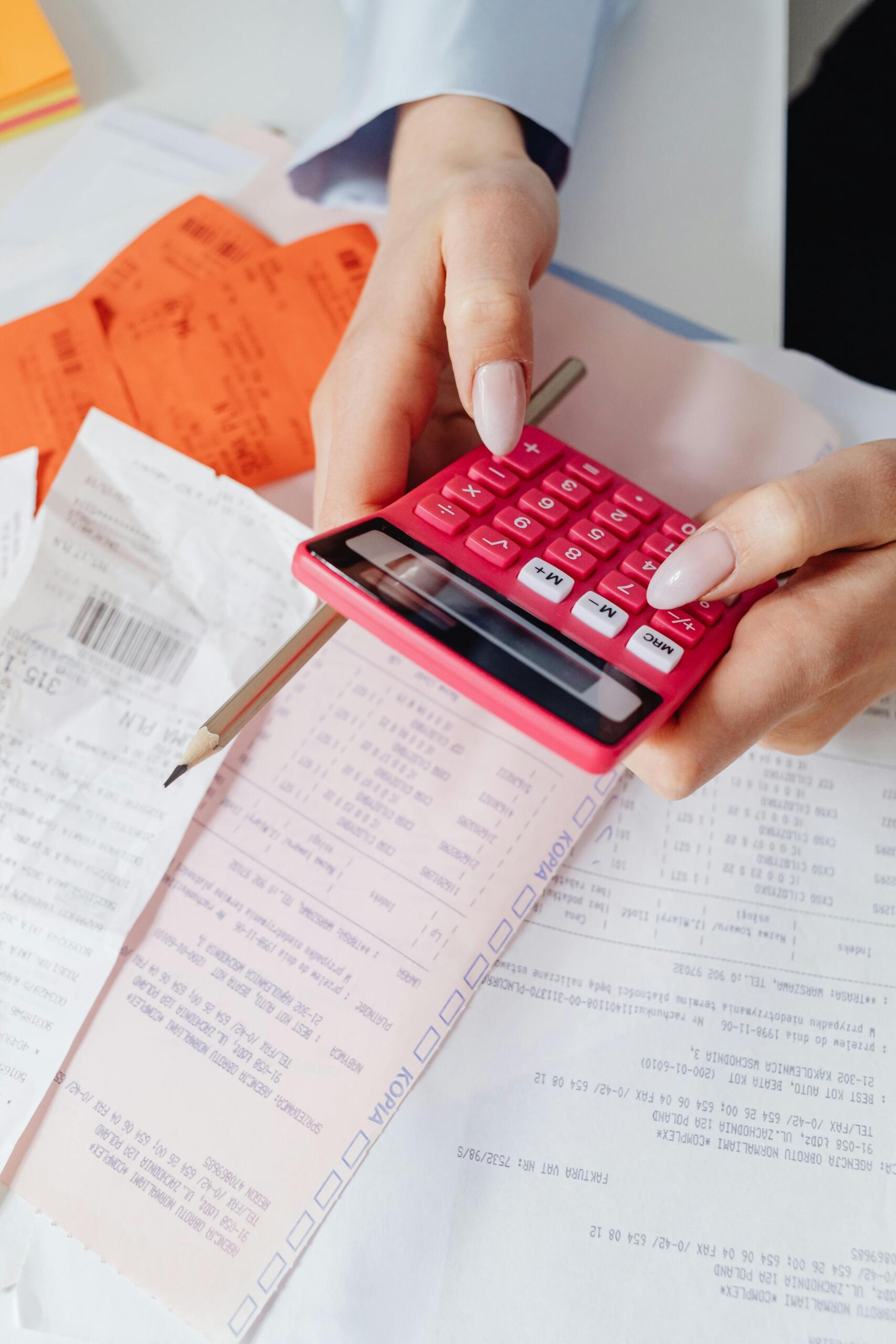 Manos femeninas usando calculadora rosa para sumar facturas y recibos sobre un escritorio, ideal para gestionar finanzas como profesor particular freelance.