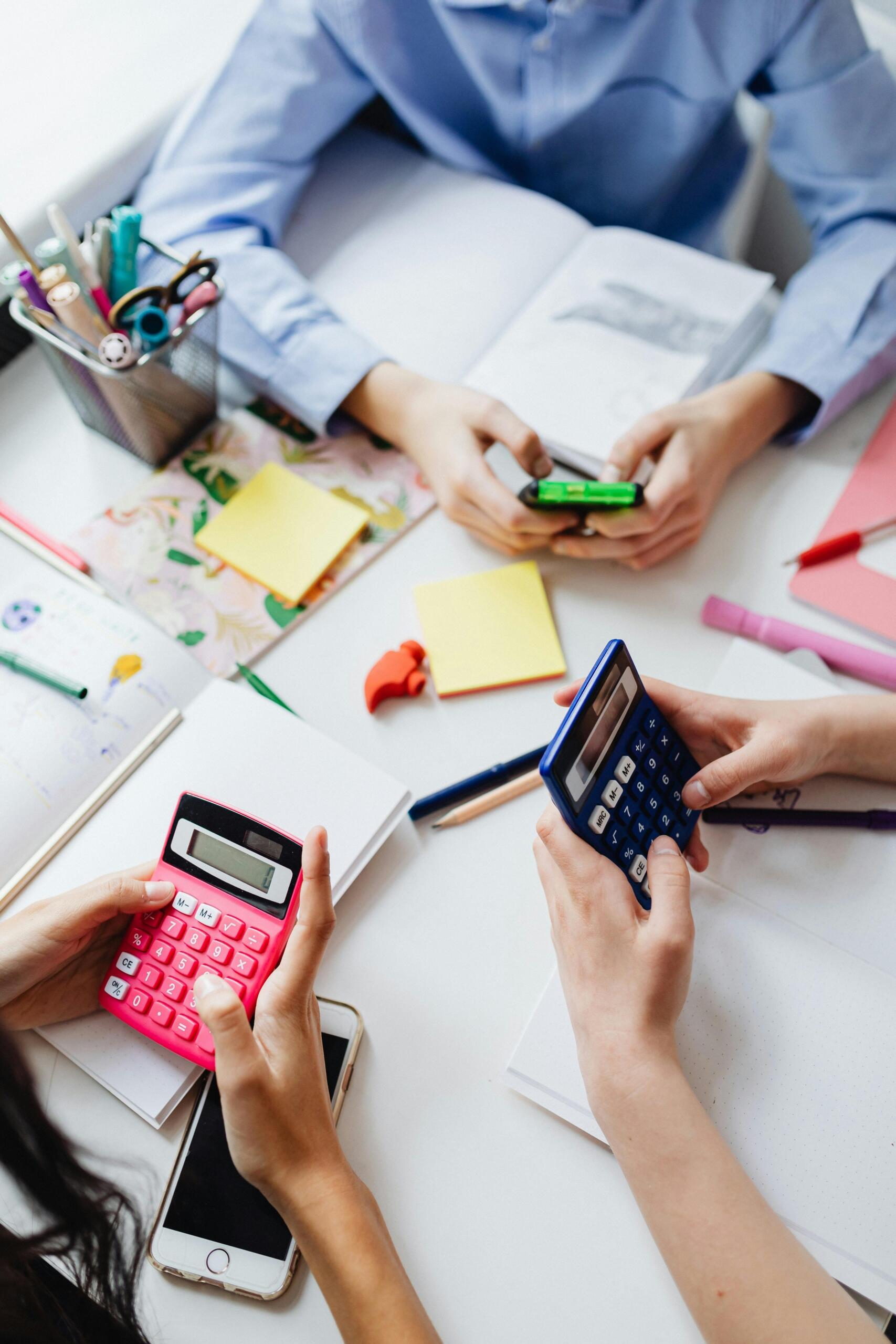 Vista cenital de dos estudiantes haciendo cálculos con calculadoras rosa y azul durante sesión de estudio en grupo con cuadernos y notas adhesivas.
