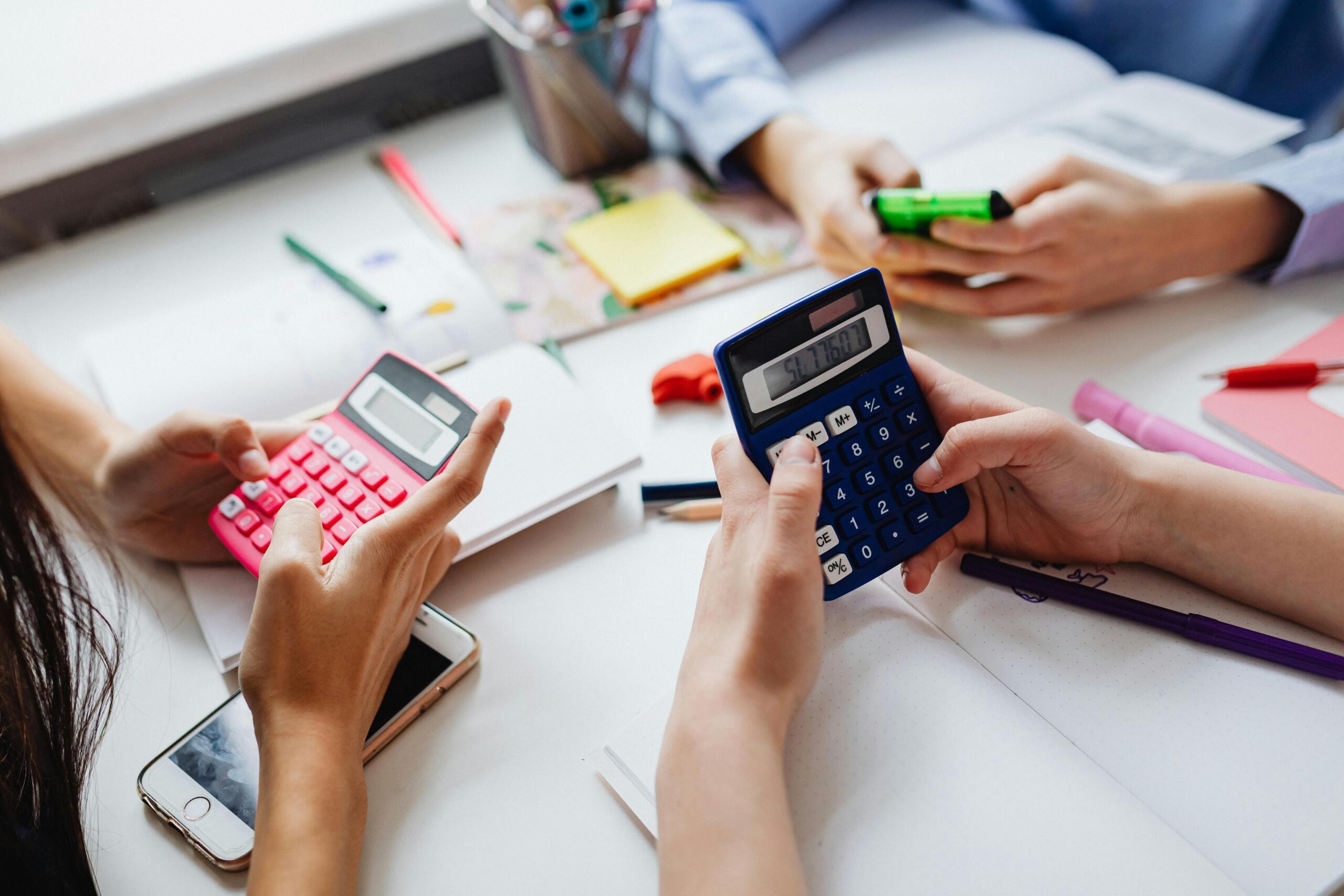 Manos sosteniendo calculadoras rosa y azul mientras hacen cuentas sobre un escritorio blanco con útiles de oficina y teléfonos móviles.