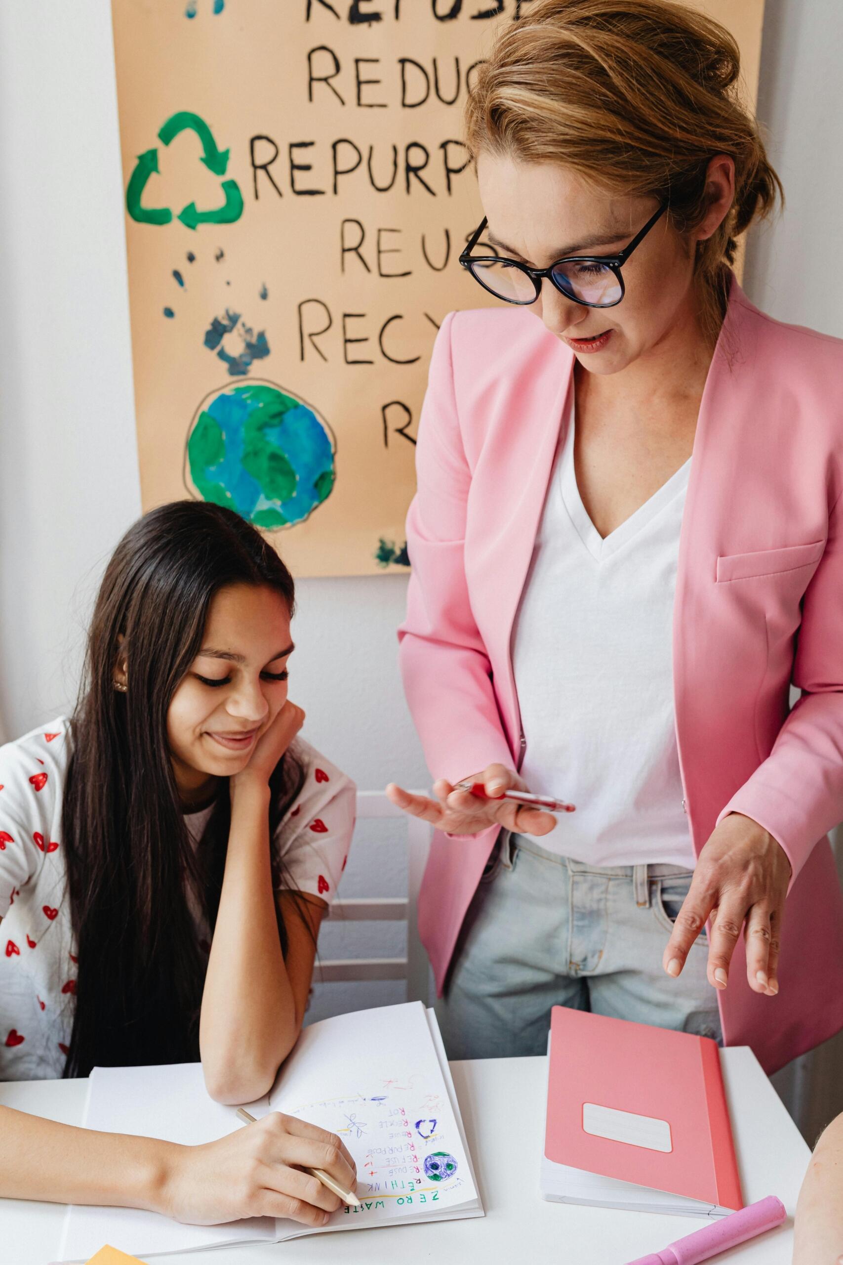 Profesora particular en blazer rosa supervisando a estudiante adolescente que escribe sobre concepto de cero residuos junto a cartel de reciclaje y sostenibilidad.