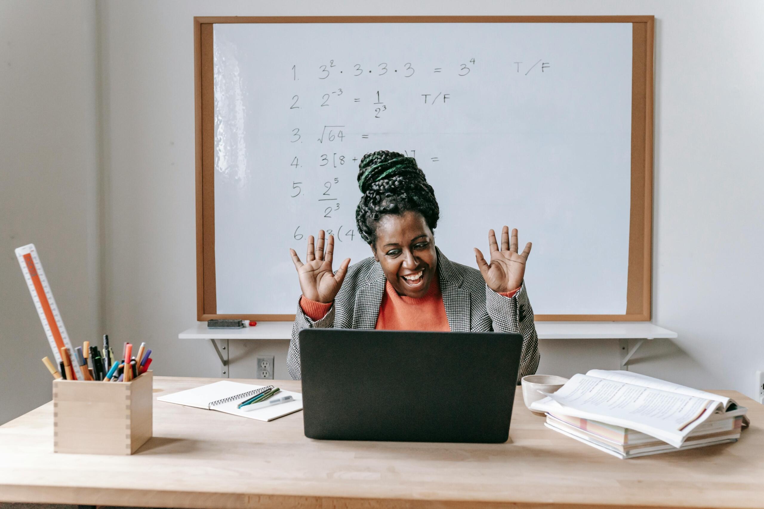 Profesora afroamericana sonriente saludando con ambas manos a sus estudiantes durante clase virtual de matemáticas con pizarra de potencias y raíces cuadradas detrás.