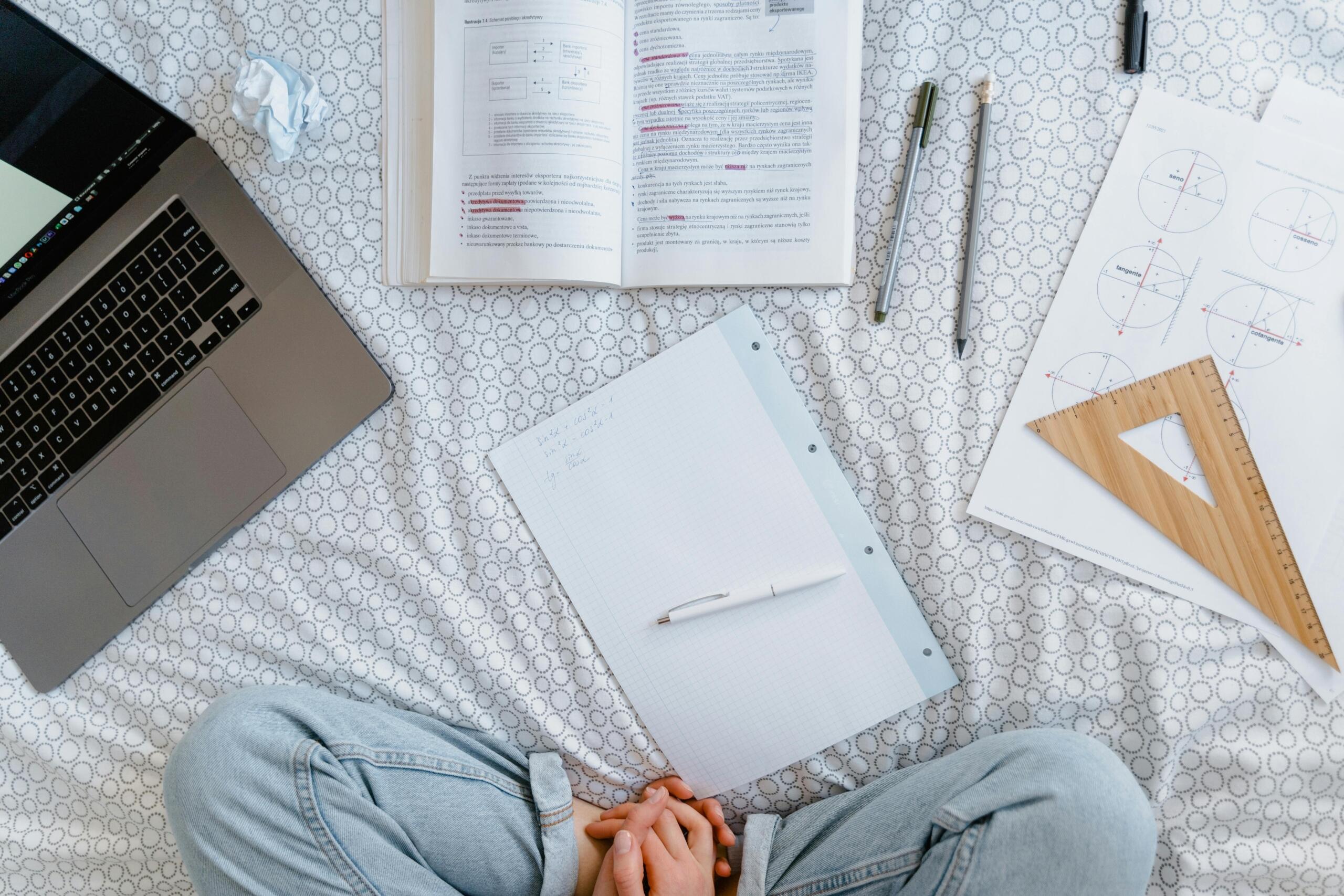 Vista superior de estudiante sentado en la cama tomando apuntes de trigonometría con laptop, libro de texto, escuadra de madera y fórmulas matemáticas.