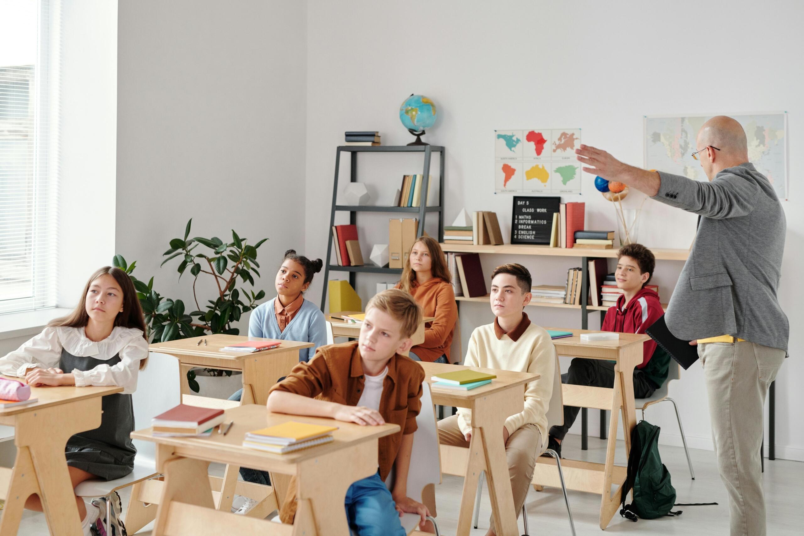 Profesor calvo señalando mapa mundial mientras imparte clase de geografía a estudiantes adolescentes diversos sentados en pupitres de un aula moderna y luminosa.