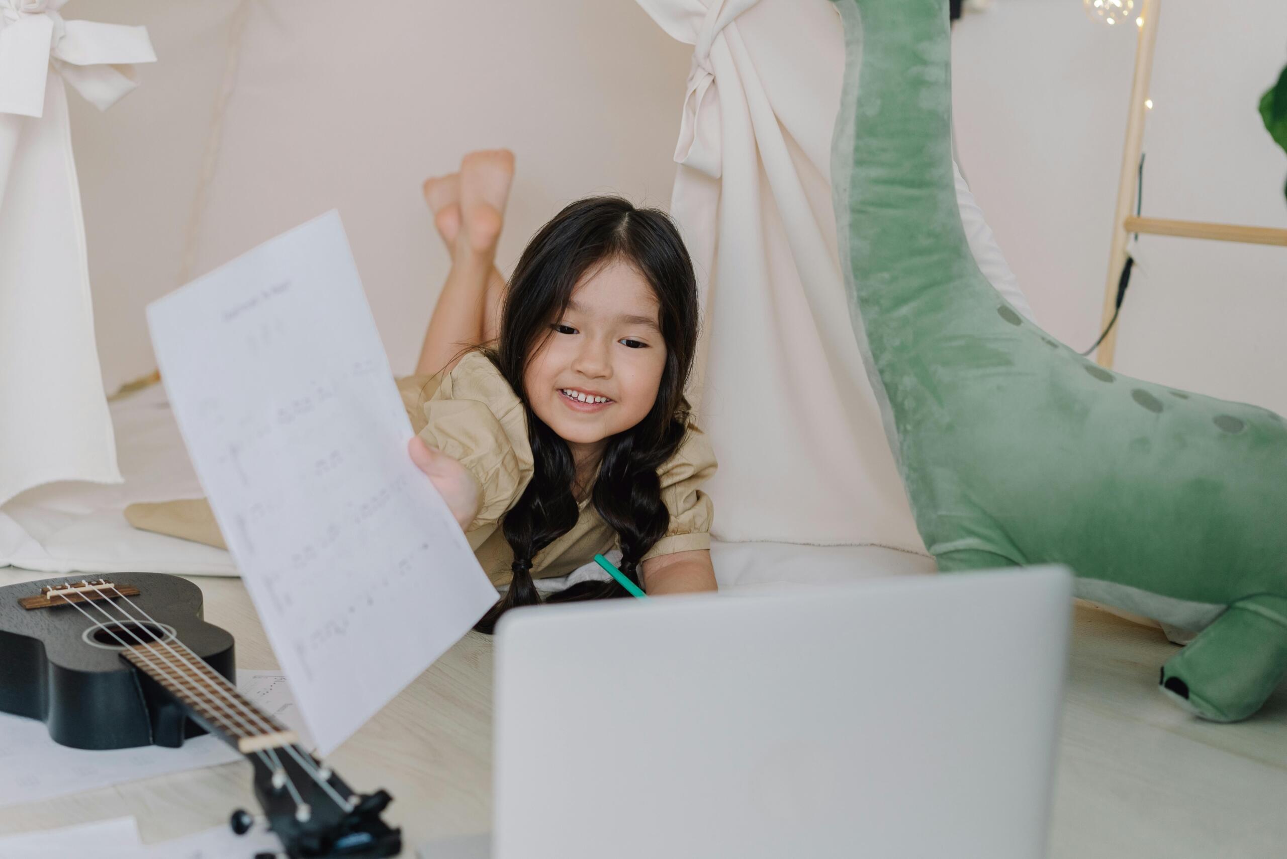 Niña asiática sonriente acostada en su carpa de juegos mostrando partitura musical frente a laptop durante clase virtual de música con ukelele a su lado.