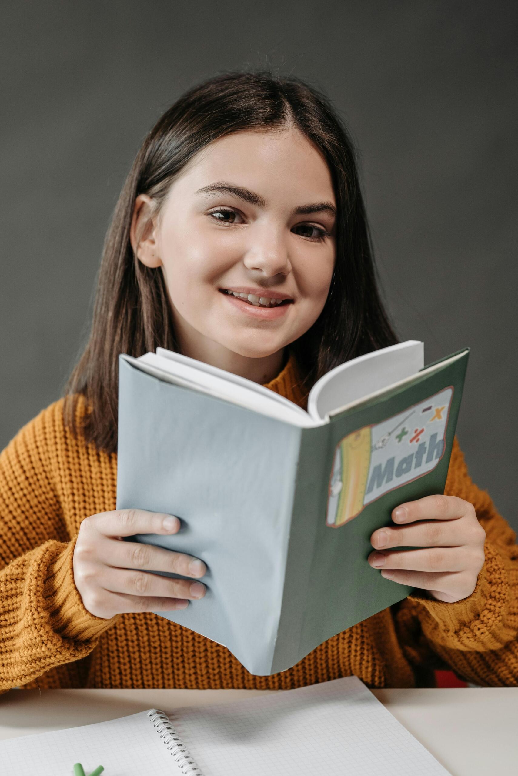 Estudiante adolescente sonriente con suéter naranja sosteniendo libro de matemáticas, mostrando entusiasmo por aprender la materia desde una actitud positiva.