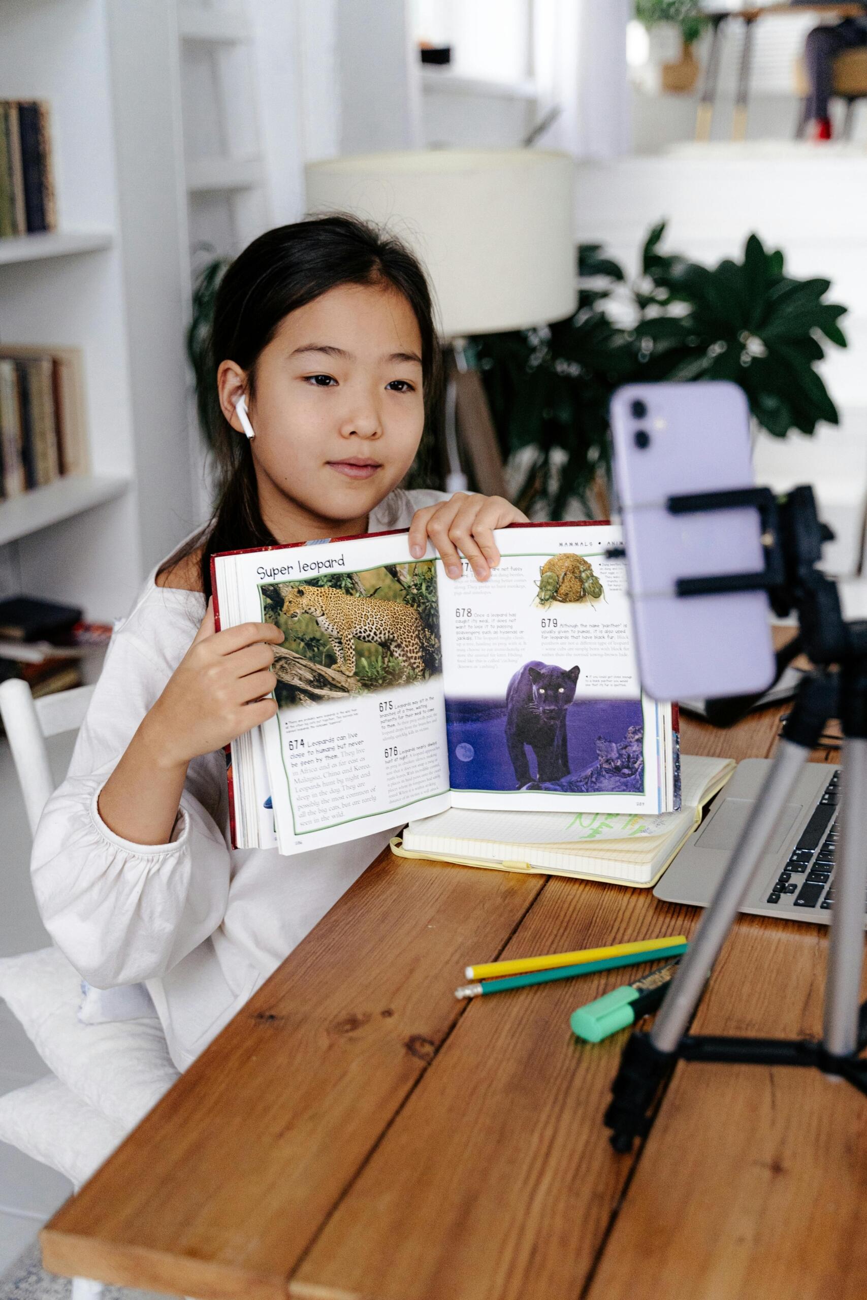 Estudiante asiática mostrando libro educativo sobre leopardos a la cámara durante clase virtual de ciencias naturales con celular en trípode y laptop.