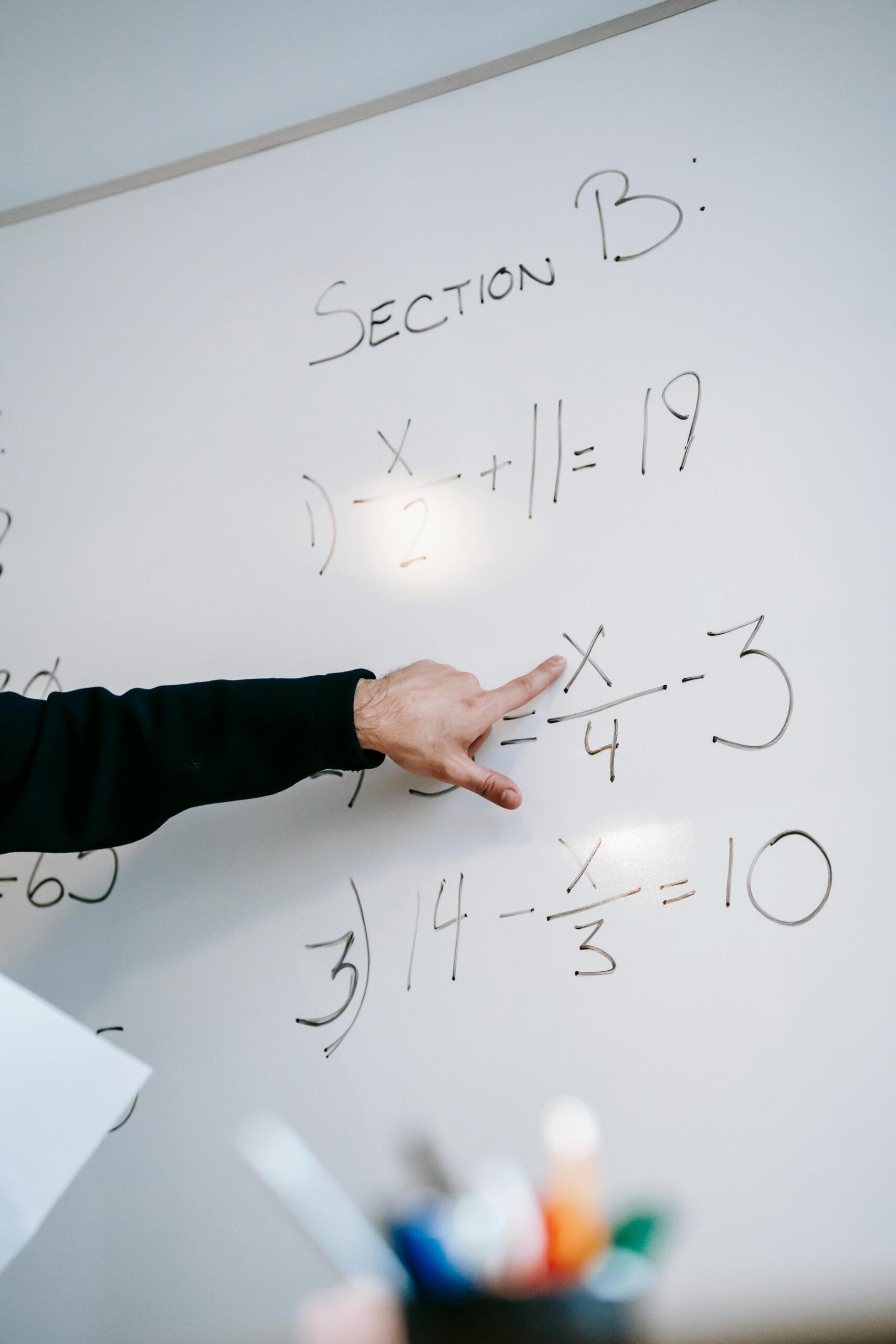 Mano de profesor señalando ecuación algebraica con fracciones en pizarra blanca durante clase de matemáticas, mostrando la sección B de un examen de álgebra.