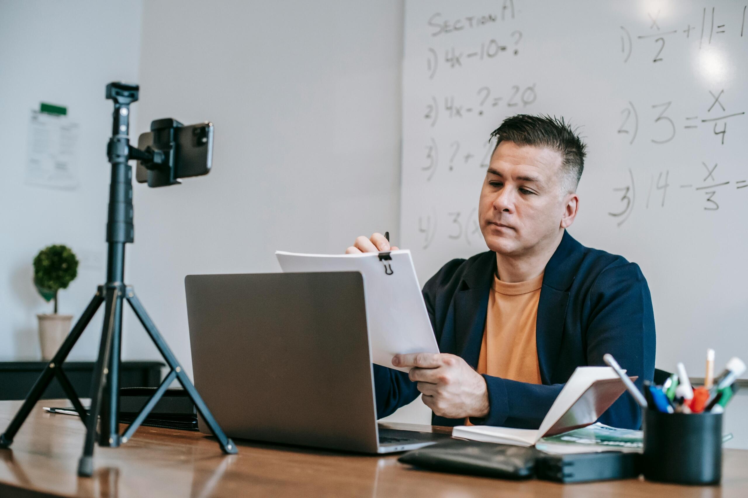 Profesor de matemáticas revisando material didáctico frente a laptop y celular en trípode durante grabación de clase online con ecuaciones algebraicas en pizarra.