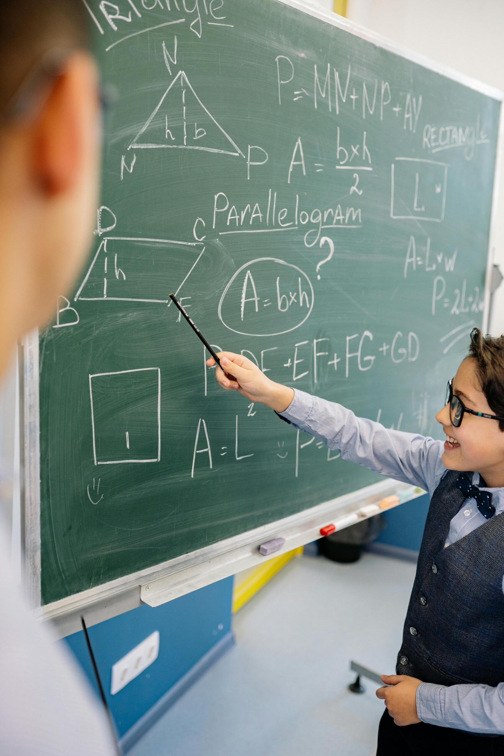 Estudiante con lentes y corbatín señalando con puntero las fórmulas de área de triángulos, paralelogramos y rectángulos en pizarra verde durante clase de geometría.
