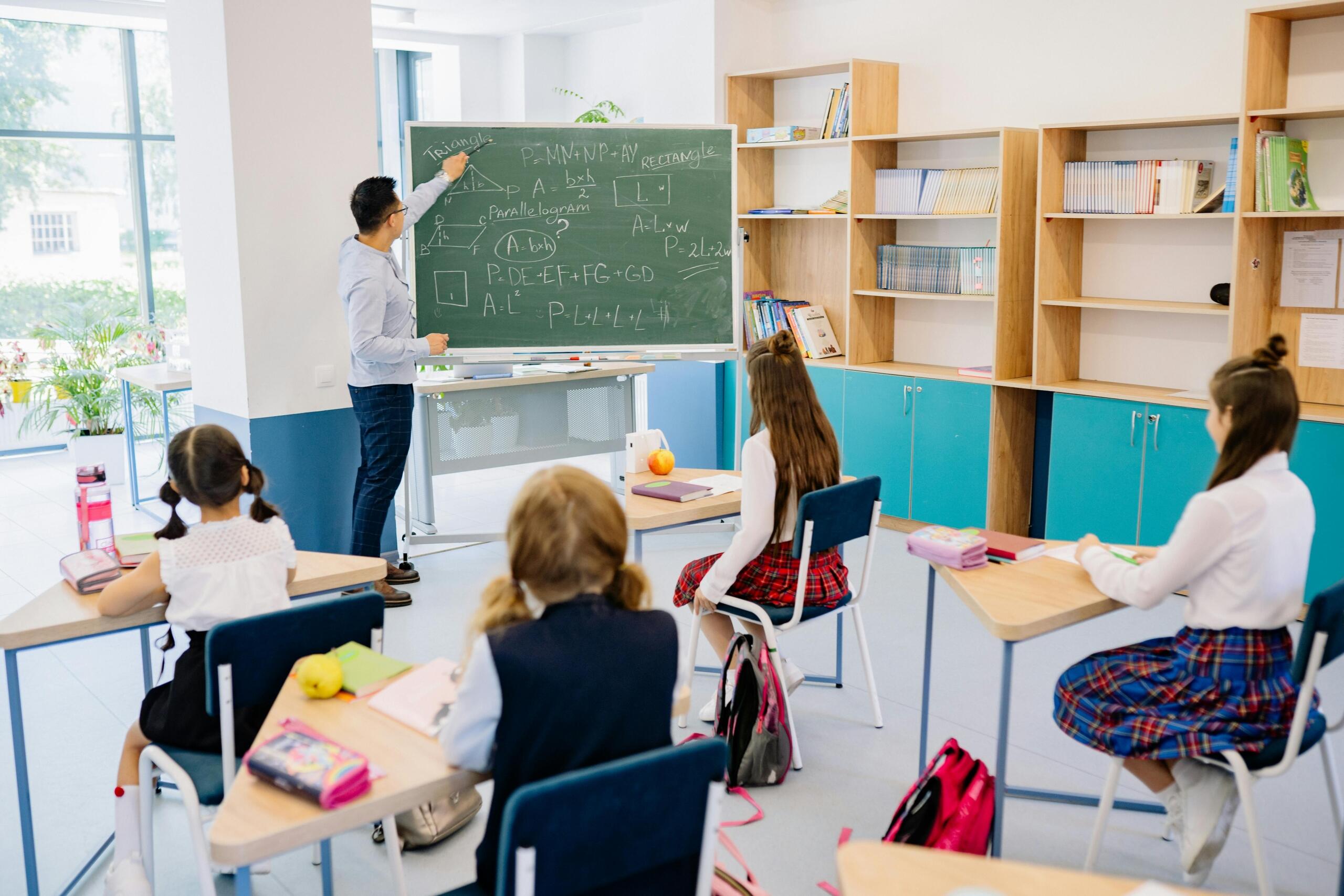 Profesor explicando fórmulas de geometría con triángulos, rectángulos y paralelogramos en pizarra a estudiantes de primaria sentadas en aula moderna y luminosa.