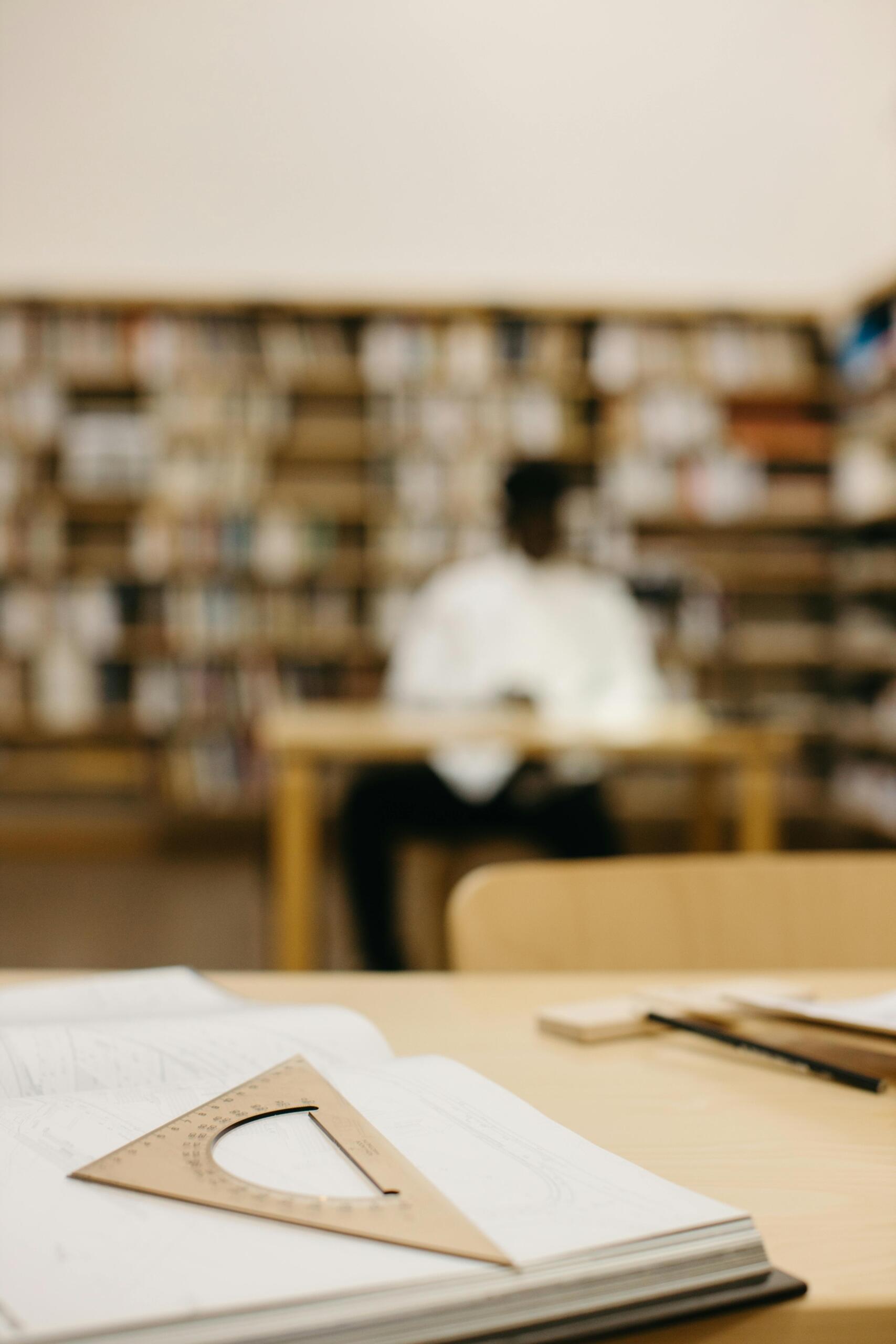 Escuadra de madera sobre cuaderno abierto en biblioteca con estudiante estudiando al fondo entre estantes de libros, ambiente ideal para repasar matemáticas en silencio.
