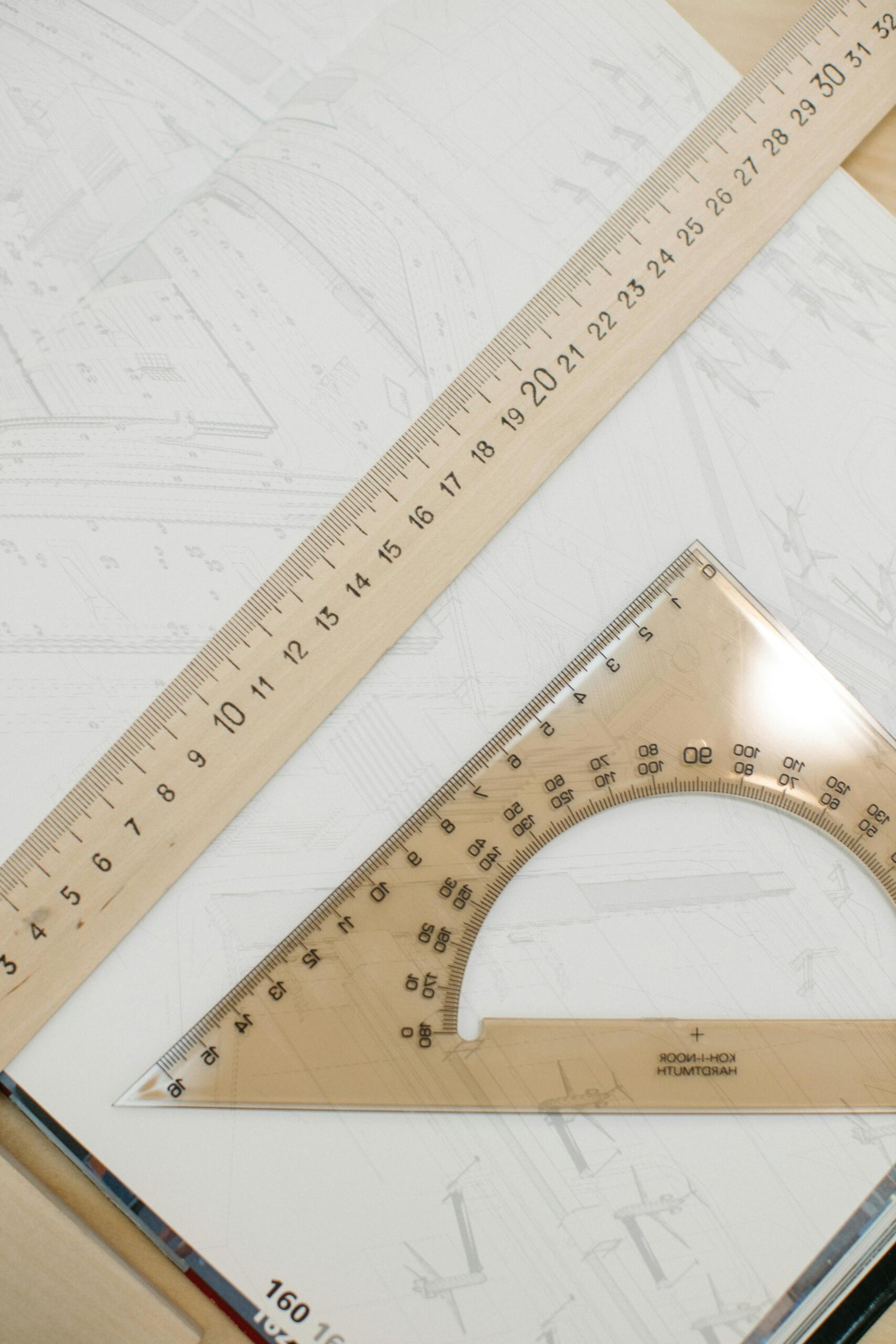 Vista cenital de regla larga de madera y escuadra triangular transparente sobre planos arquitectónicos, herramientas esenciales para el dibujo técnico y la geometría.
