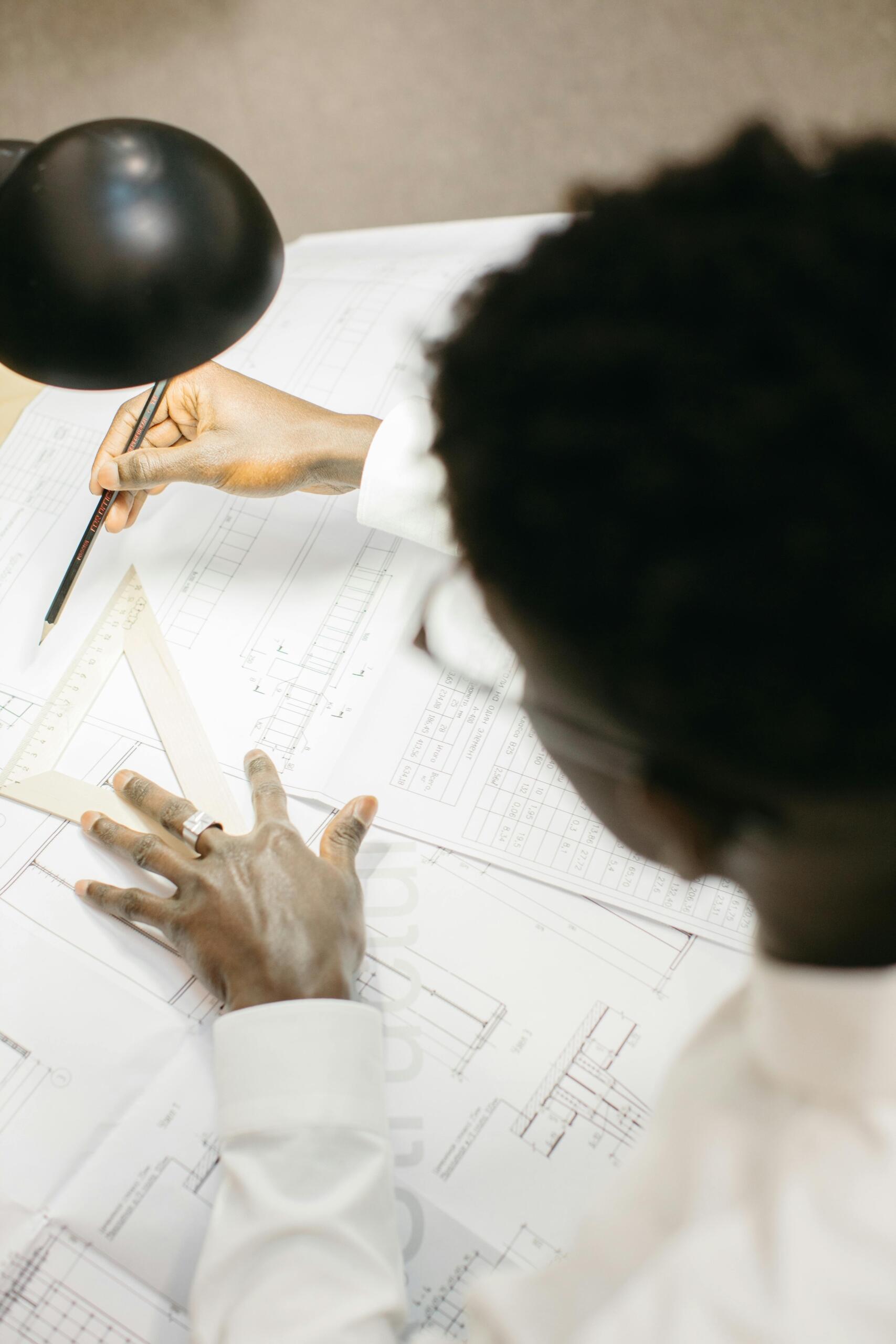Estudiante afroamericano tomando medidas con escuadra y lápiz sobre planos de ingeniería bajo lámpara de escritorio, aplicando matemáticas al diseño técnico.