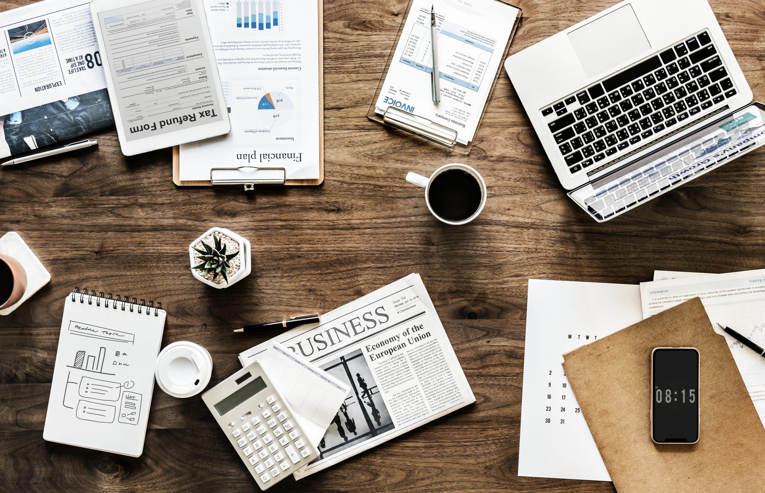mesa de trabajo con reportes económicos, gráficos y diarios sobre economia junto a una laptop