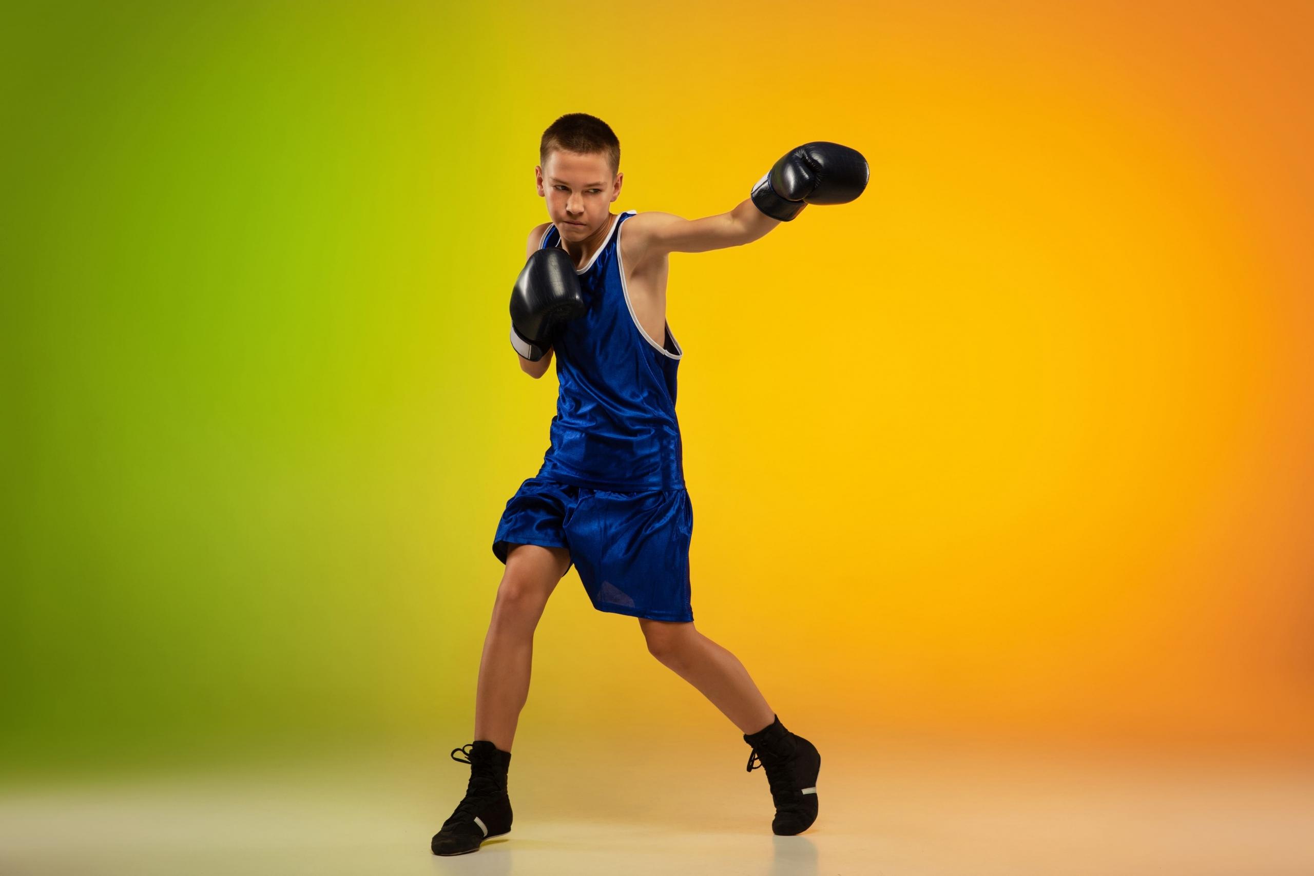 niño adolescente con ropa deportiva y guantes de box lanzando un puño al aire y fondo de color verde naranja