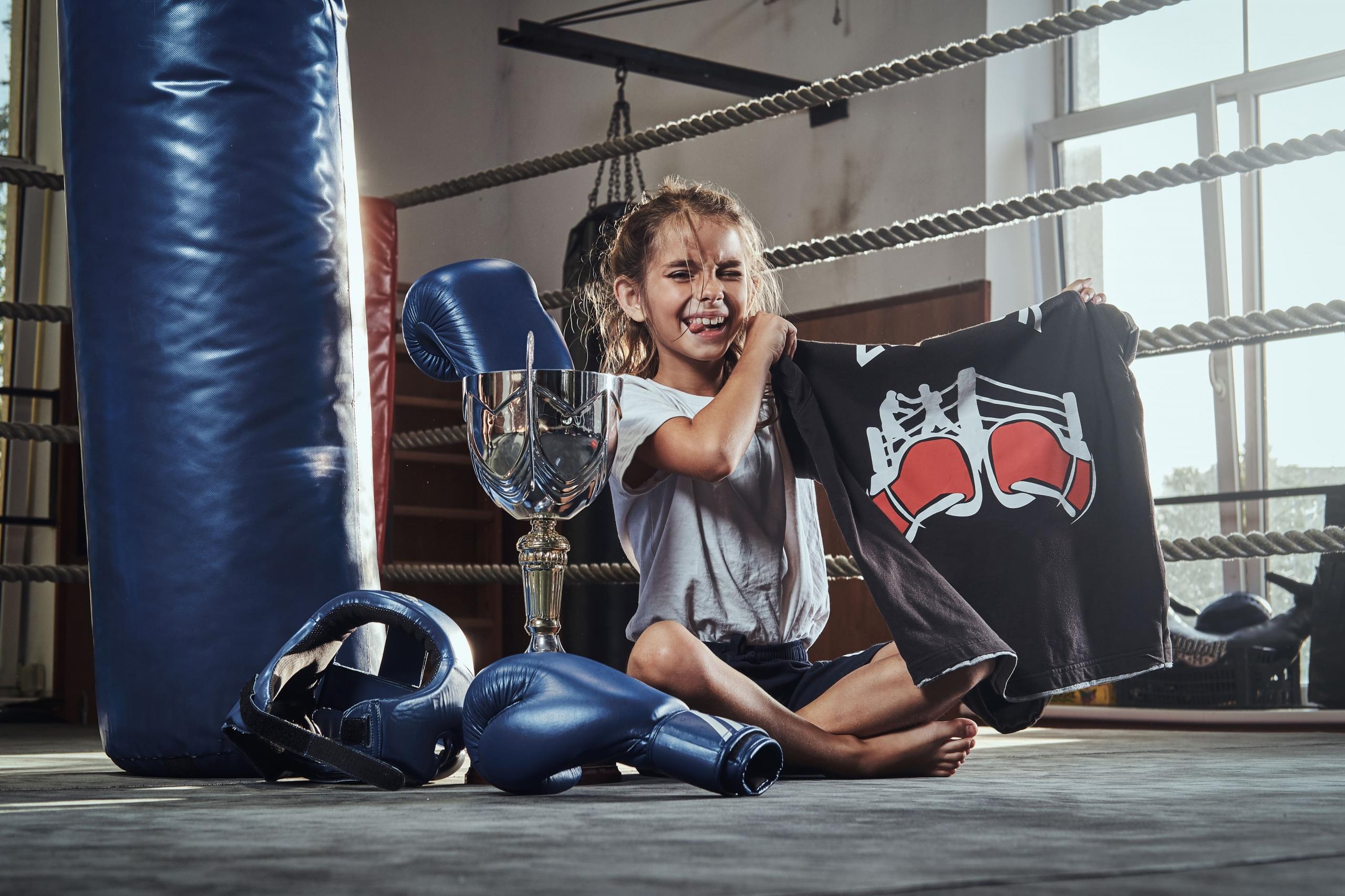 niña sentada en un ring sosteniendo un polo de box, guantes y un trofeo