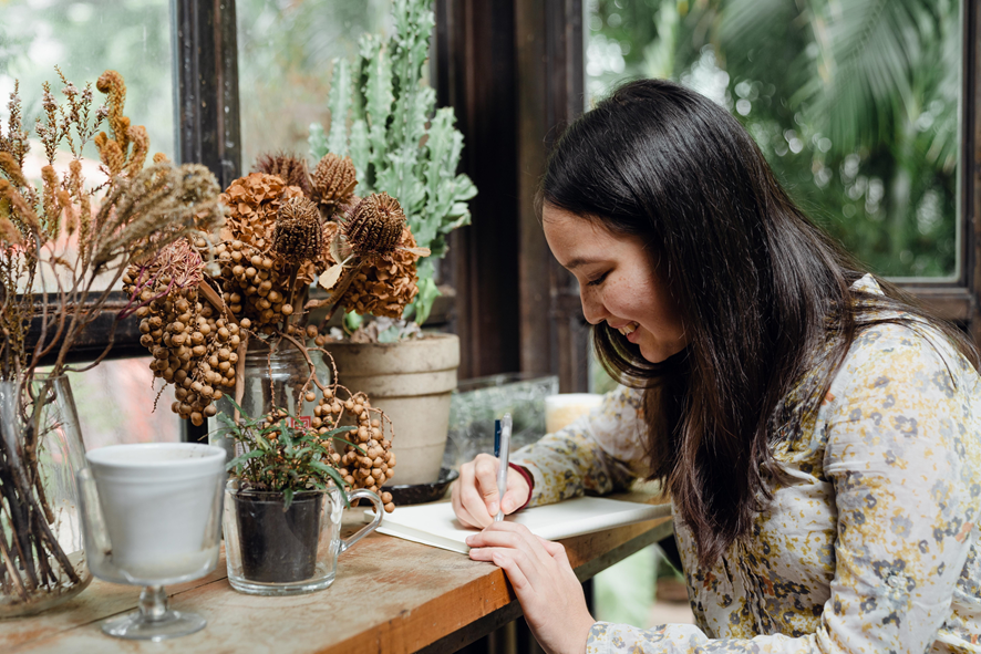 chica escribiendo con un lapicero en unas hojas, sobre una mesa llena de arreglos florales.