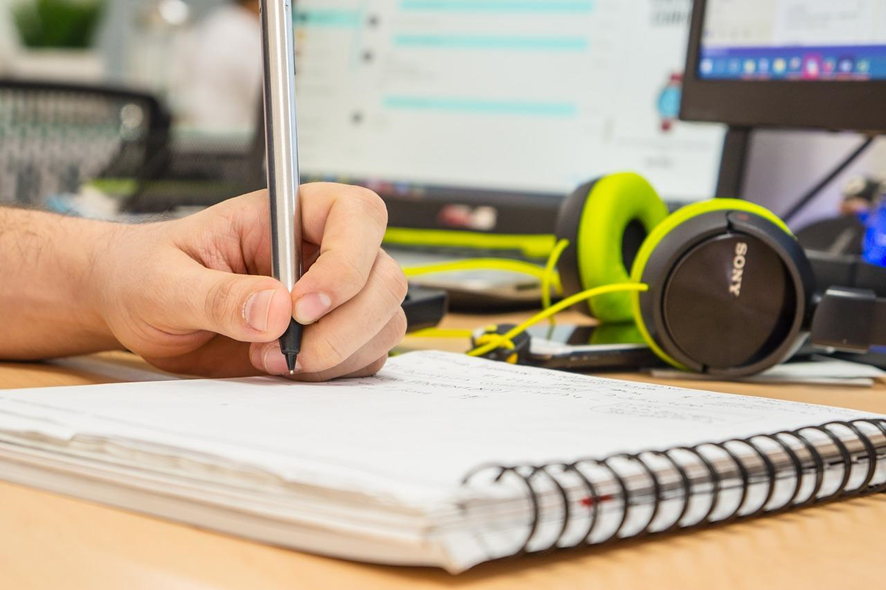 mano escribiendo sobre una libreta con audífonos y computadora sobre la mesa