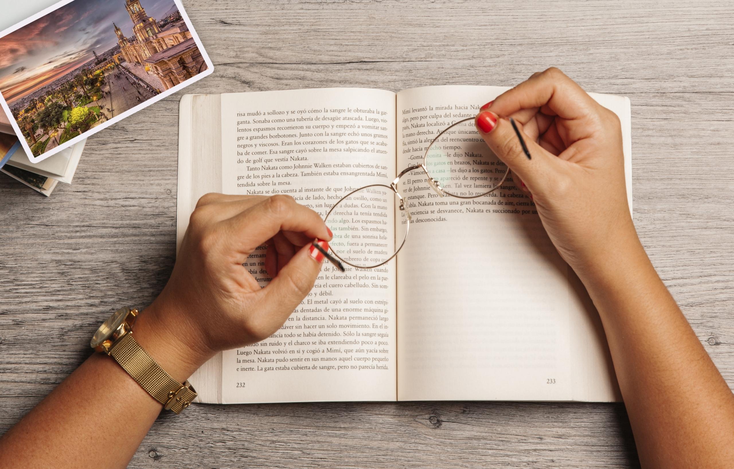 manos de mujer sosteniendo unos lentes sobre un libro de español y una postal de Arequipa sobre la mesa