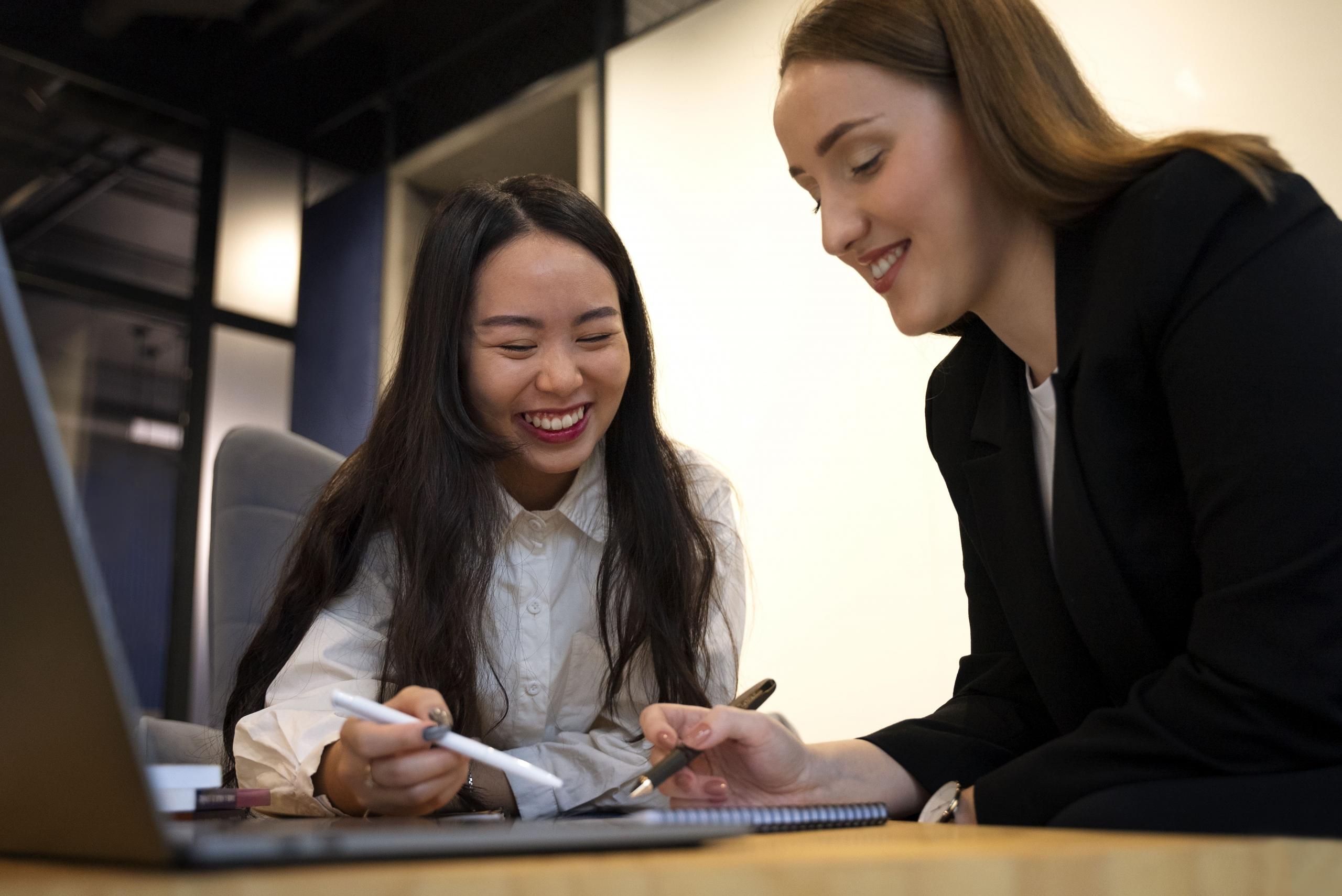 dos mujeres sonrientes revisando sus anotaciones frente a una laptop