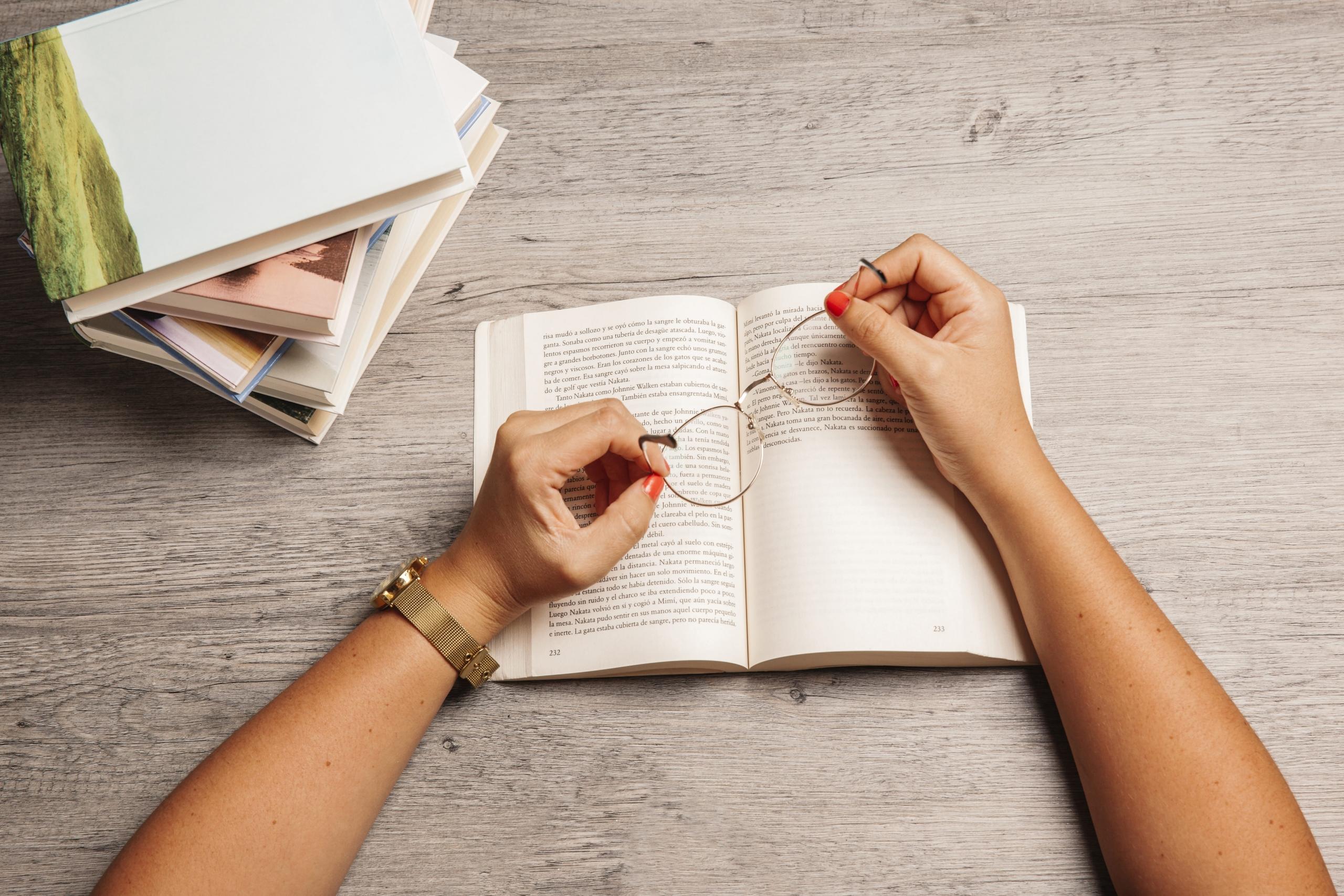 vista detalle de unas manos sosteniendo unos lentes sobre un libro abierto con paginas de un texto en español