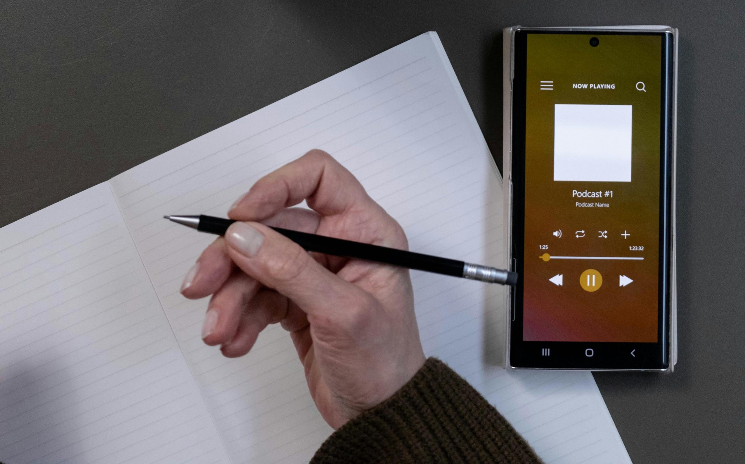mano escribiendo mientras sostiene el lapiz y hay un celular al costado reproduciendo un podcast