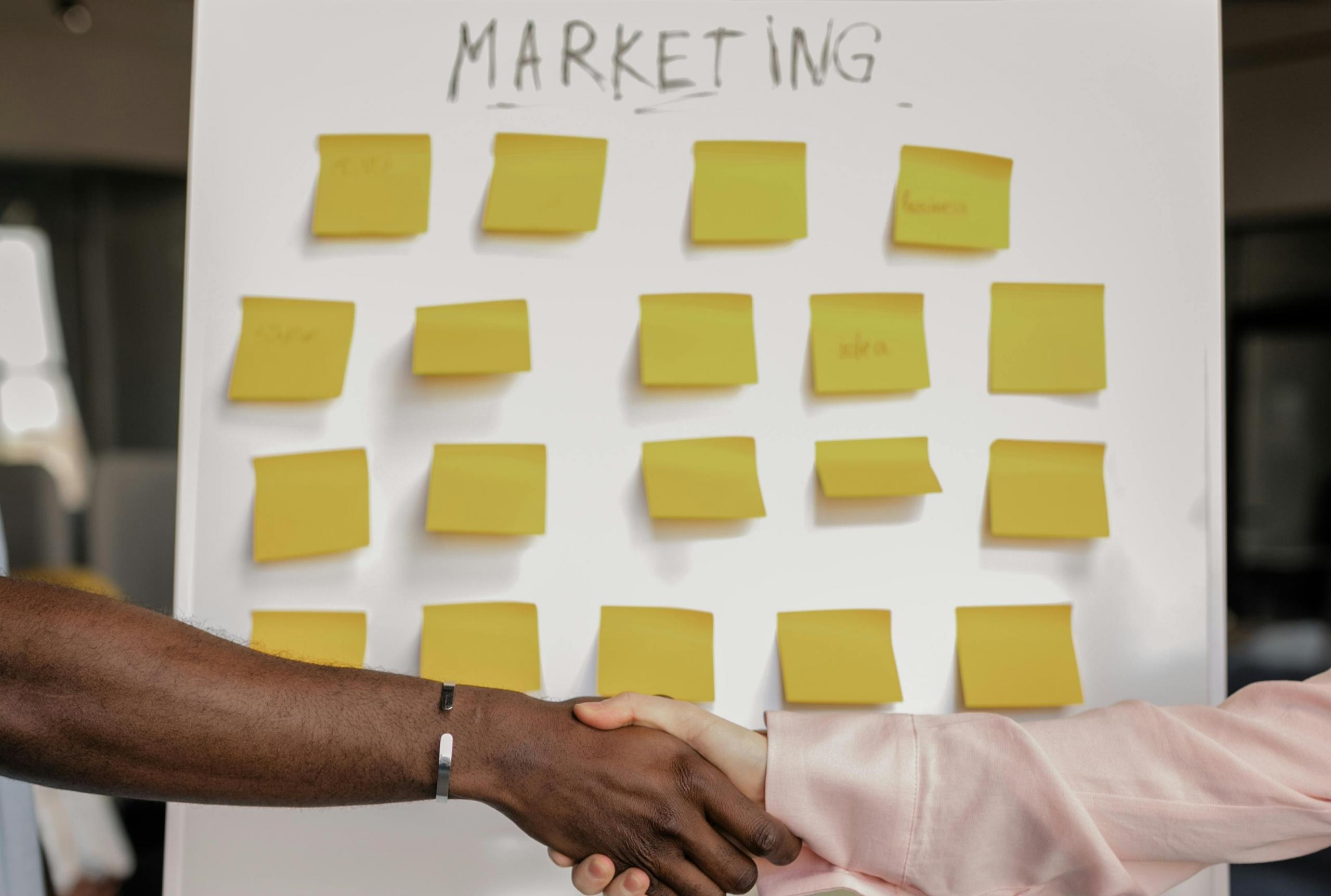 dos manos estrechandose frente a una pizarra llena de post-its y la pallabra MARKETING escrita en la parte superior