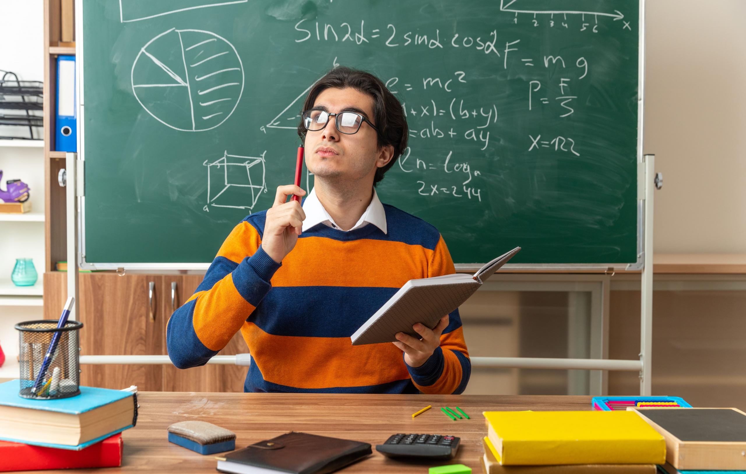 chico pensativo con lapiz y cuaderno en la mano con fondo de salon de clase y pizarra llena de matemáticas