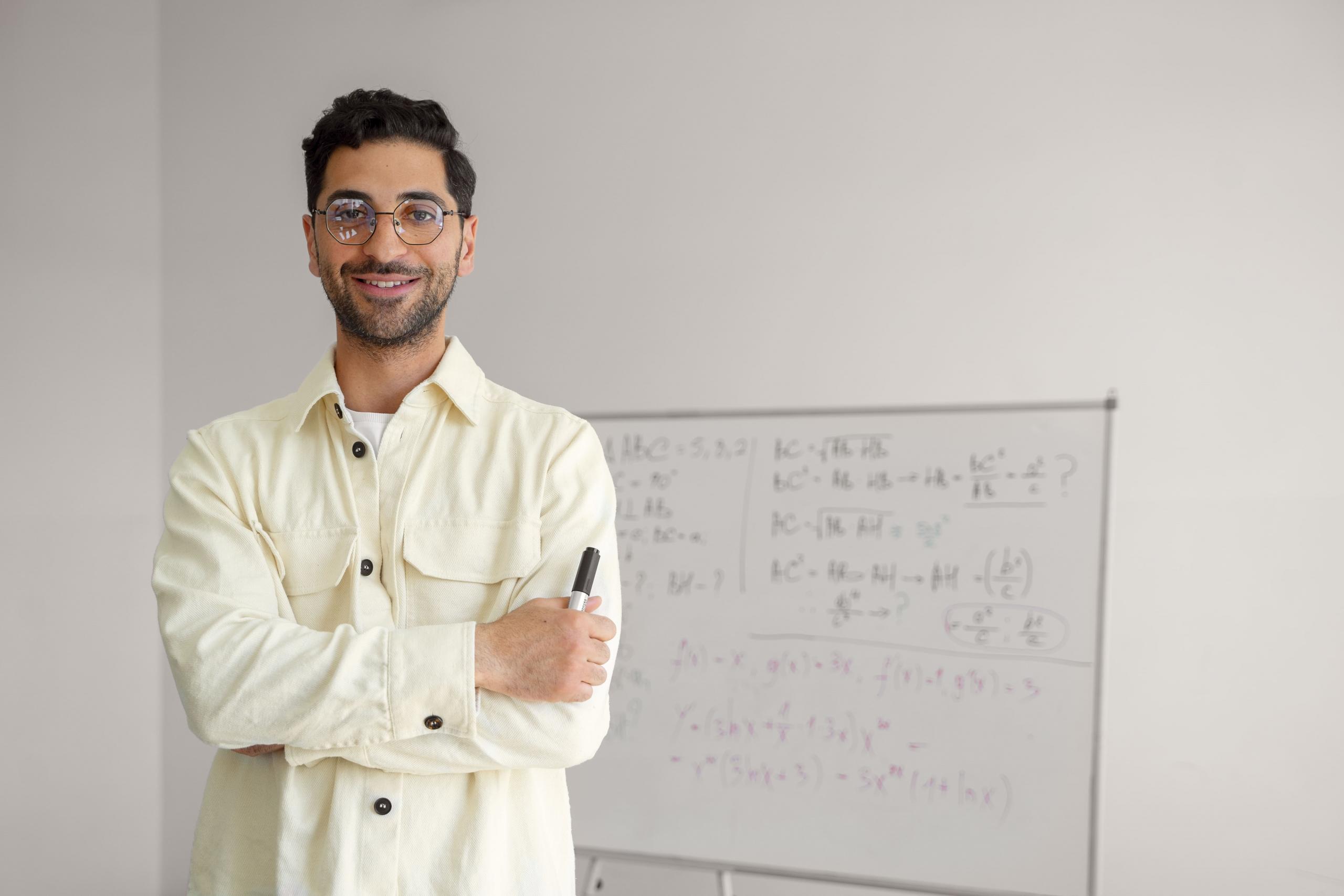 profesor sonriente sosteniendo un plumón en la mano y pizarra llena de ejercicios matemáticos de fondo
