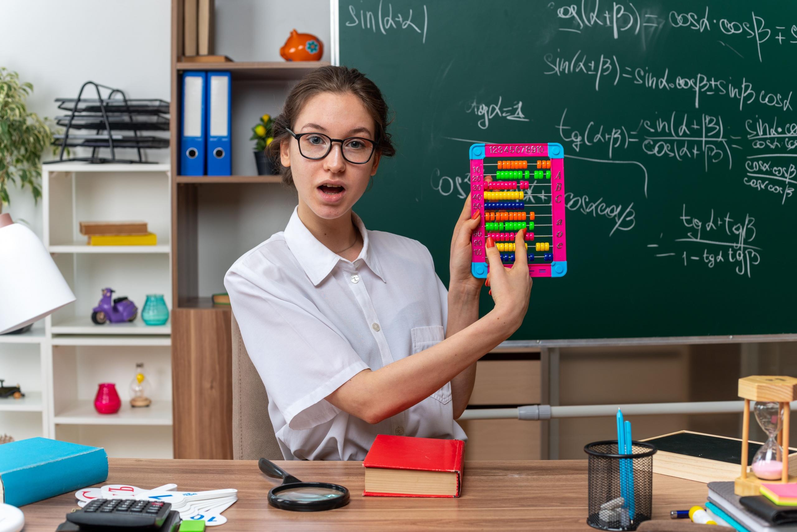 profesora sosteniendo un ábaco sorprendida con pizarrón de formulas matemáticas de fondo en un aula.