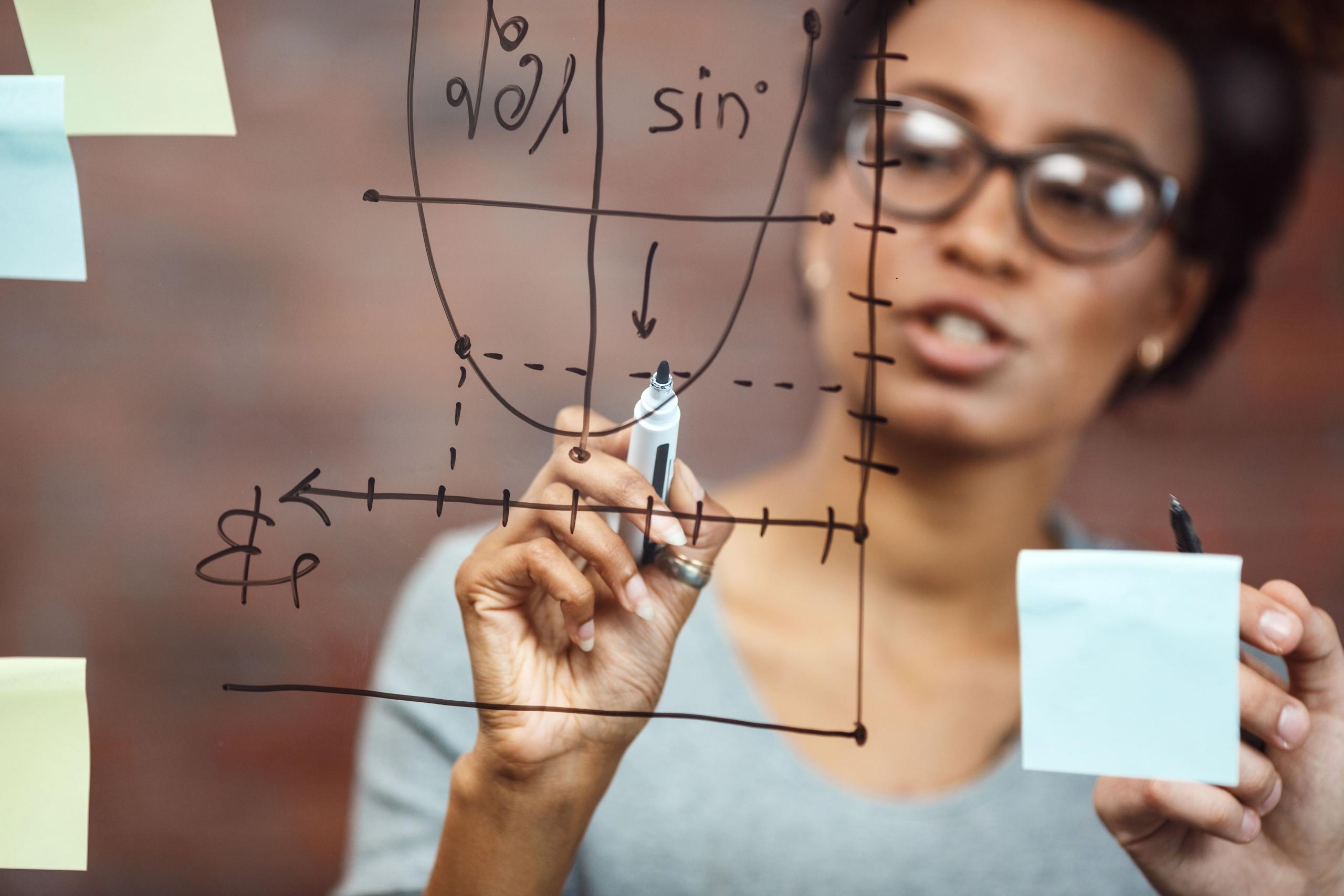 vista de un ejercicio matemático en primer plano en pizarra transparente y mujer resolviendo el problema con un plumón