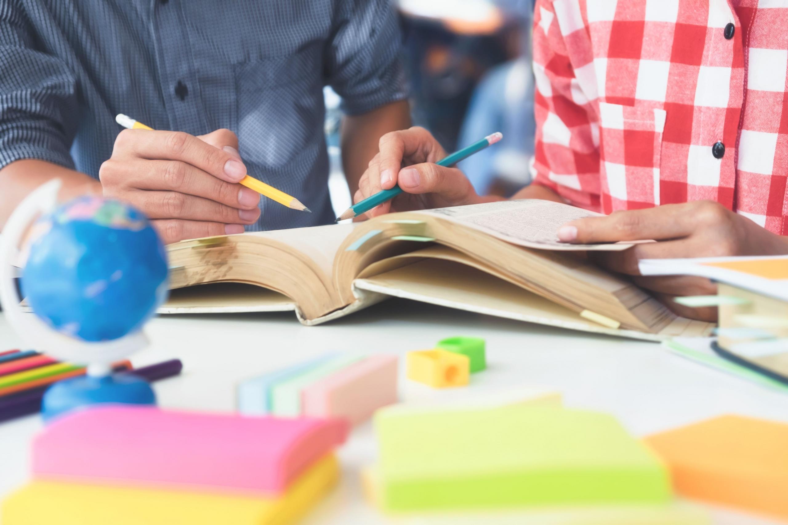 manos de dos personas con lapices y libros con anotaciones y post its alrededor