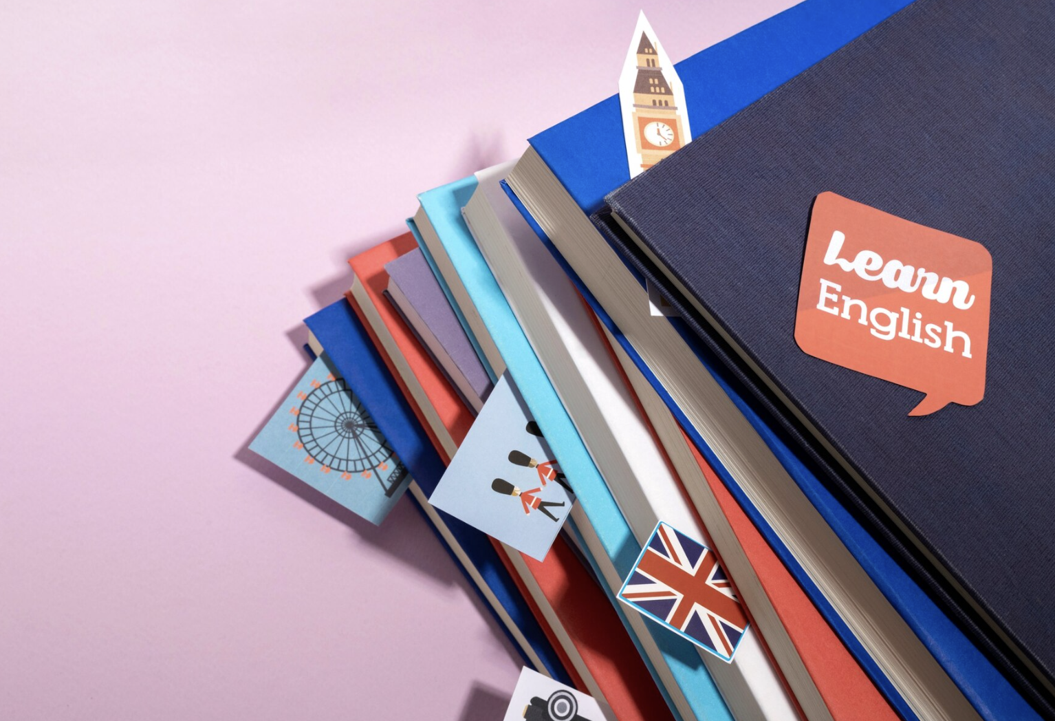 vista cenital de varios libros de ingles apilados con recortes de diversas figuritas del london eye, bandera inglesa, los soldados, big ben y el globito de LEARN ENGLISH en el libro superior