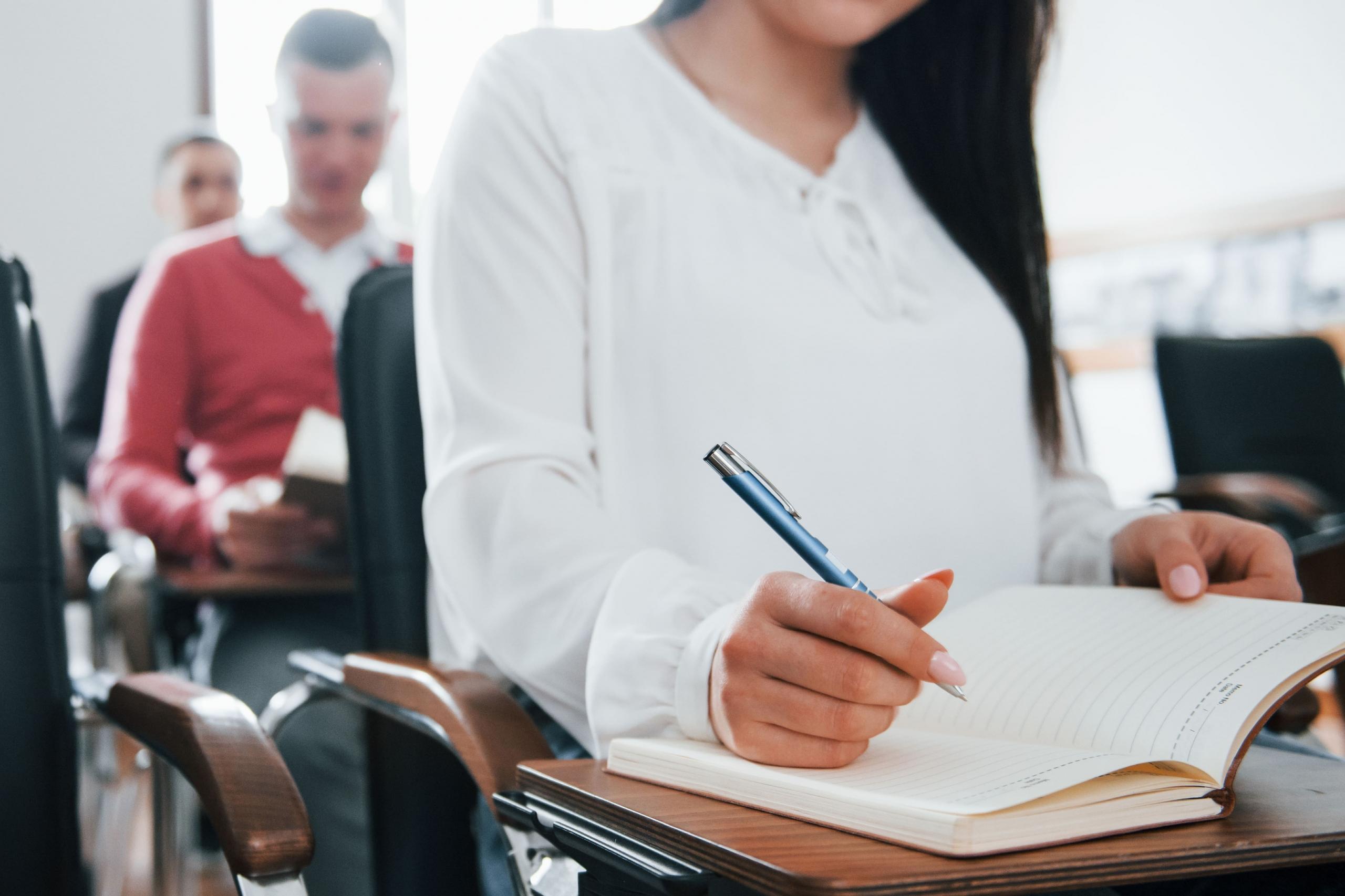 vista entrecortada de torso mujer en un escritorio con una agenda y lapicero en mano, con otros estudiantes de fondo