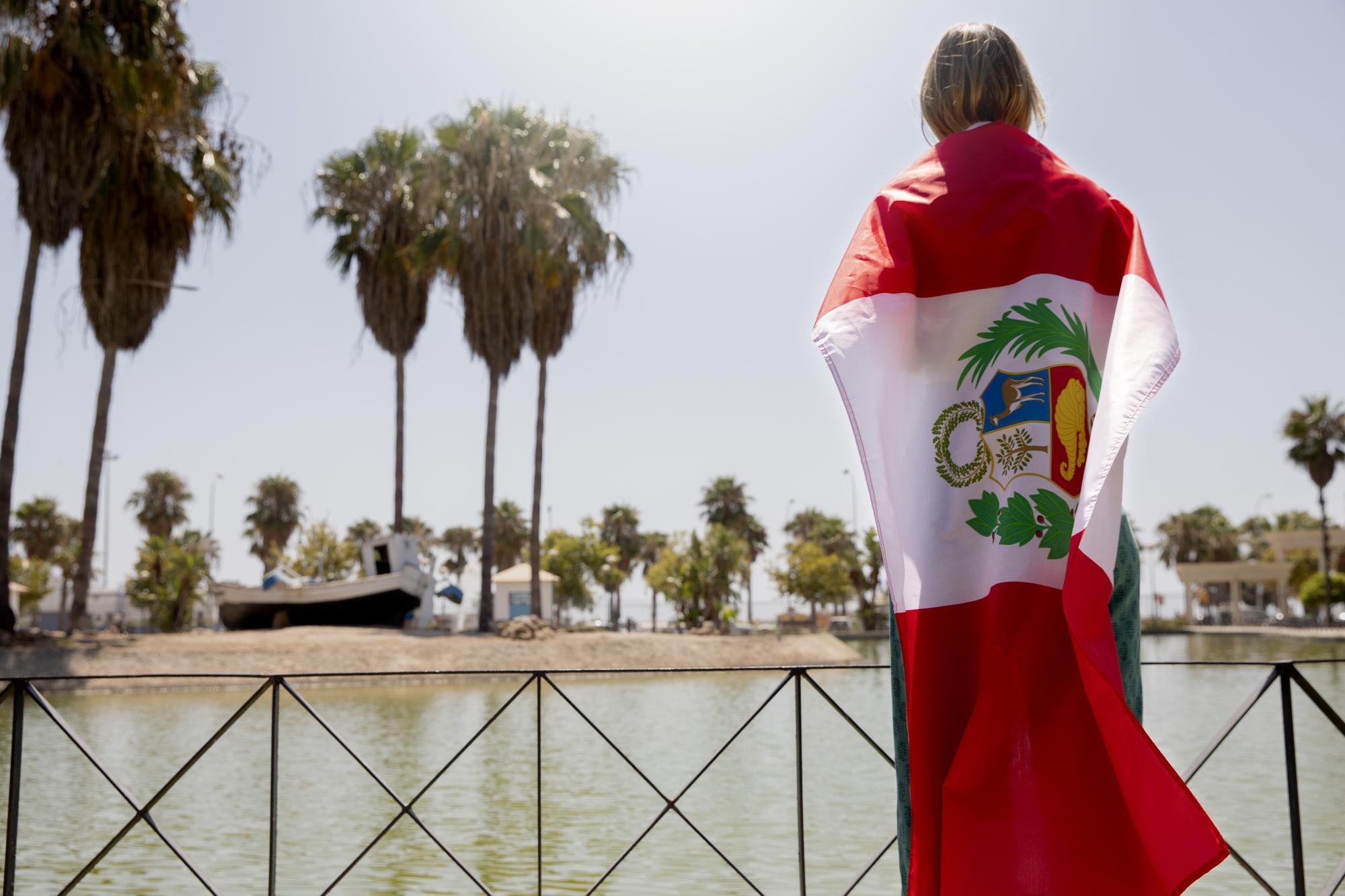 mujer parada de espaldas a la cámara con una bandera del perú cubriendo todo su cuerpo junto a una laguna con palmeras alrededor.