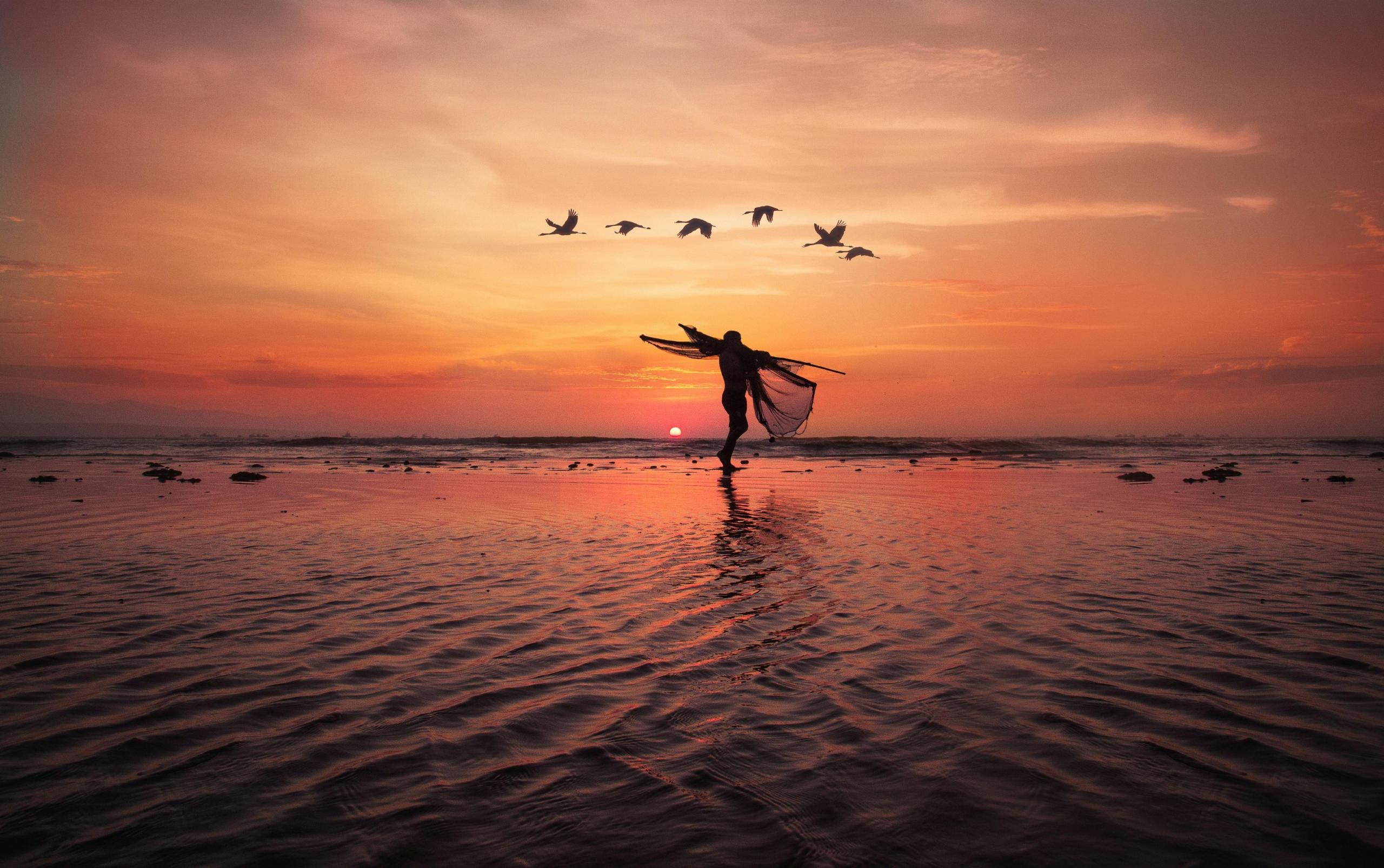 vista de un sunset con la silueta de una persona con redes de pescar a lo lejos y seis aves sobrevolando el cielo naranja