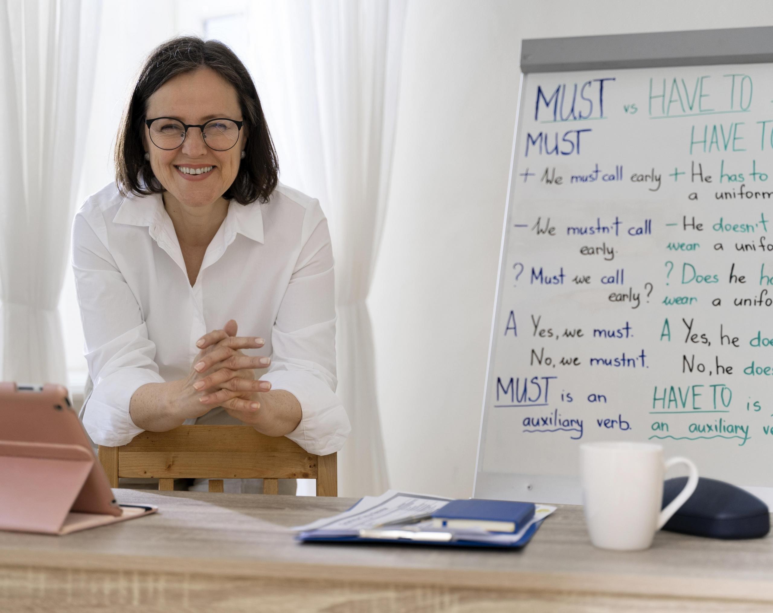 profesora sonriente mirando a la cámara junto a su pizarra llena de una leccion de inglés