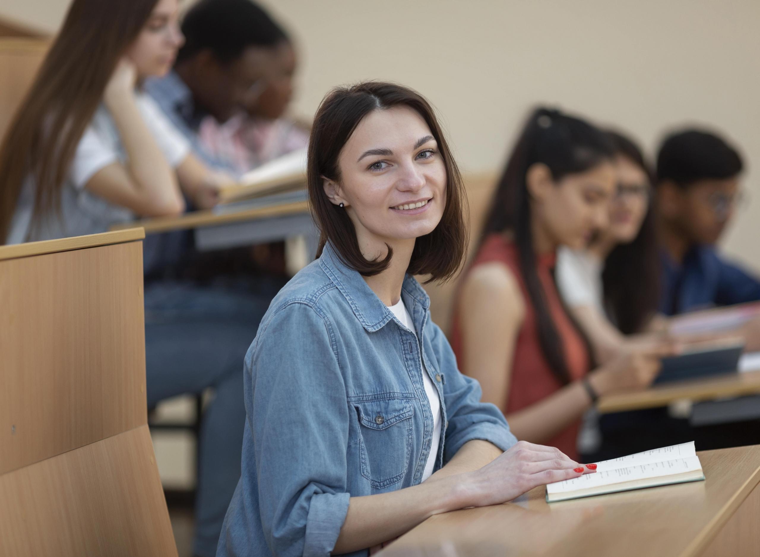 vista de chica mirando a la cámara con un libro y otros alumnos de fondo en el mismo aula