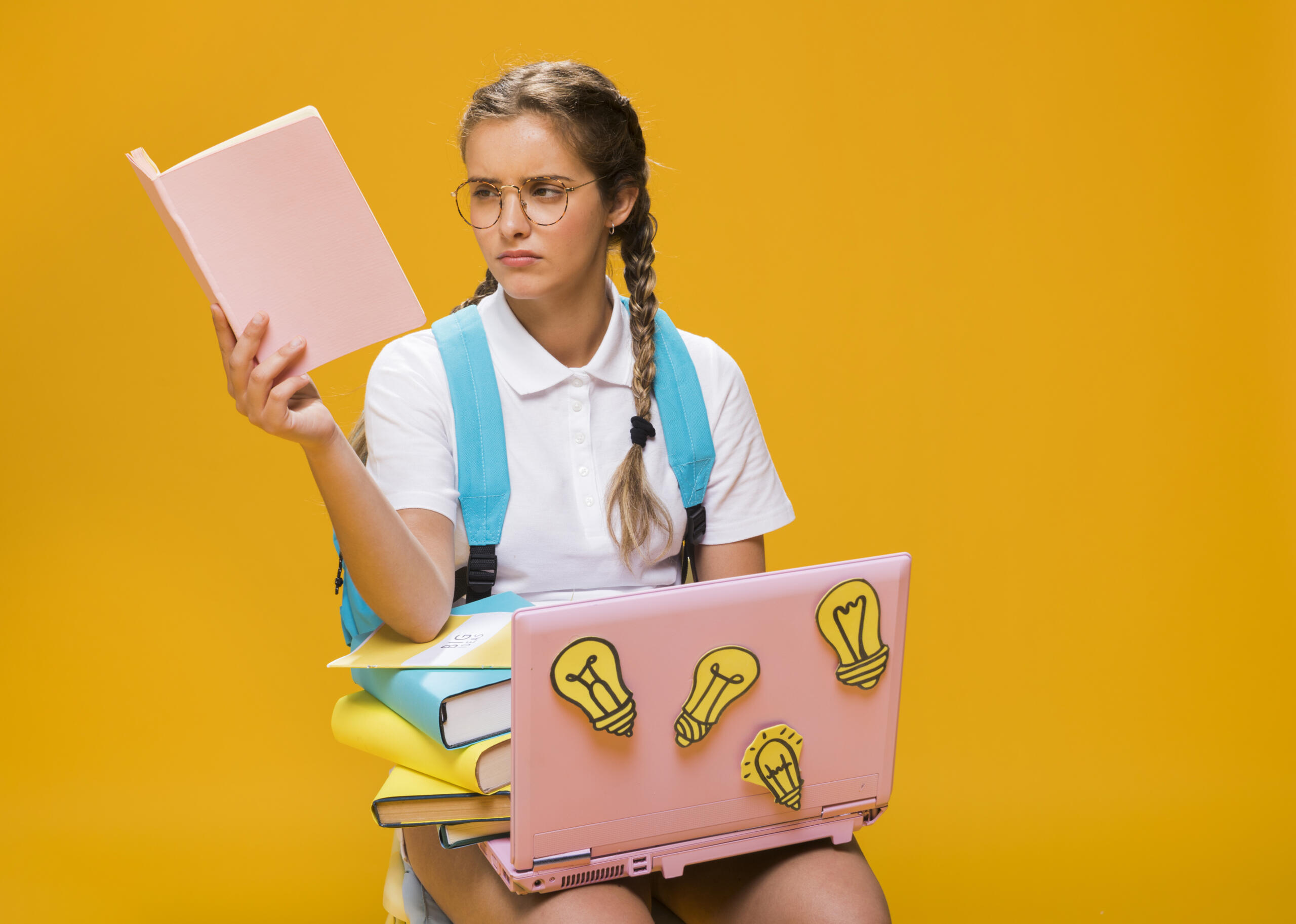 chica con un libro en mano y rostro confundido, sentada y sosteniendo libros y una laptop sobre sus piernas