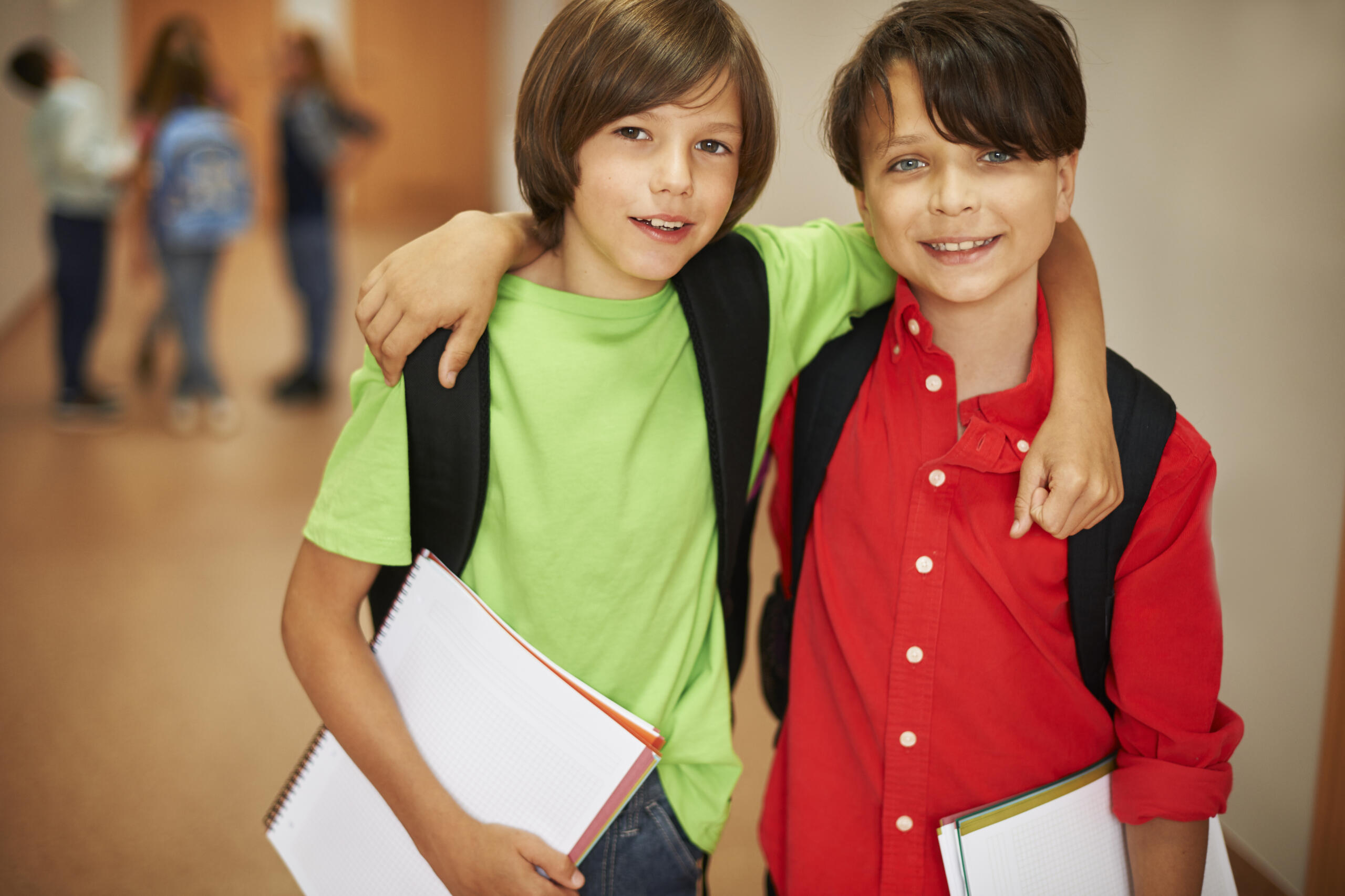 dos niños sonrientes con camisa verde y roja abrazados sosteniendo sus cuadernos y mochila en la espalda