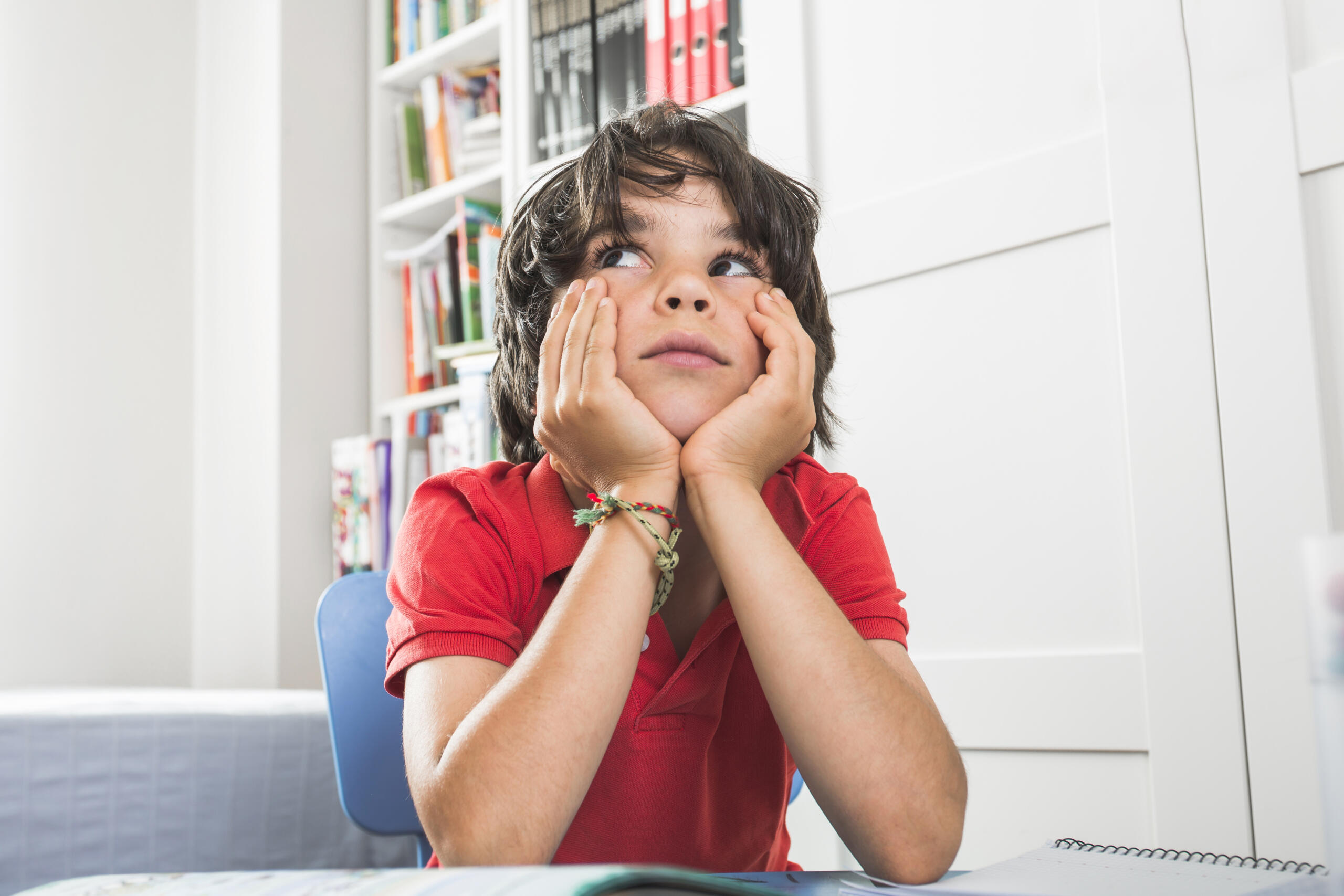 niño con polo rojo apoyado con sus dos codos en escritorio pensativo mirando hacia arriba