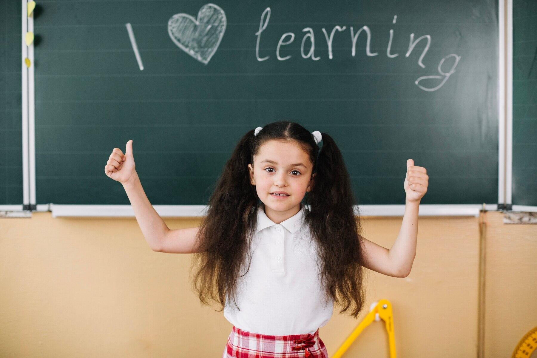 niña con los dos pulgares arriba y letras escritas en la pizarra I LOVE LEARNING