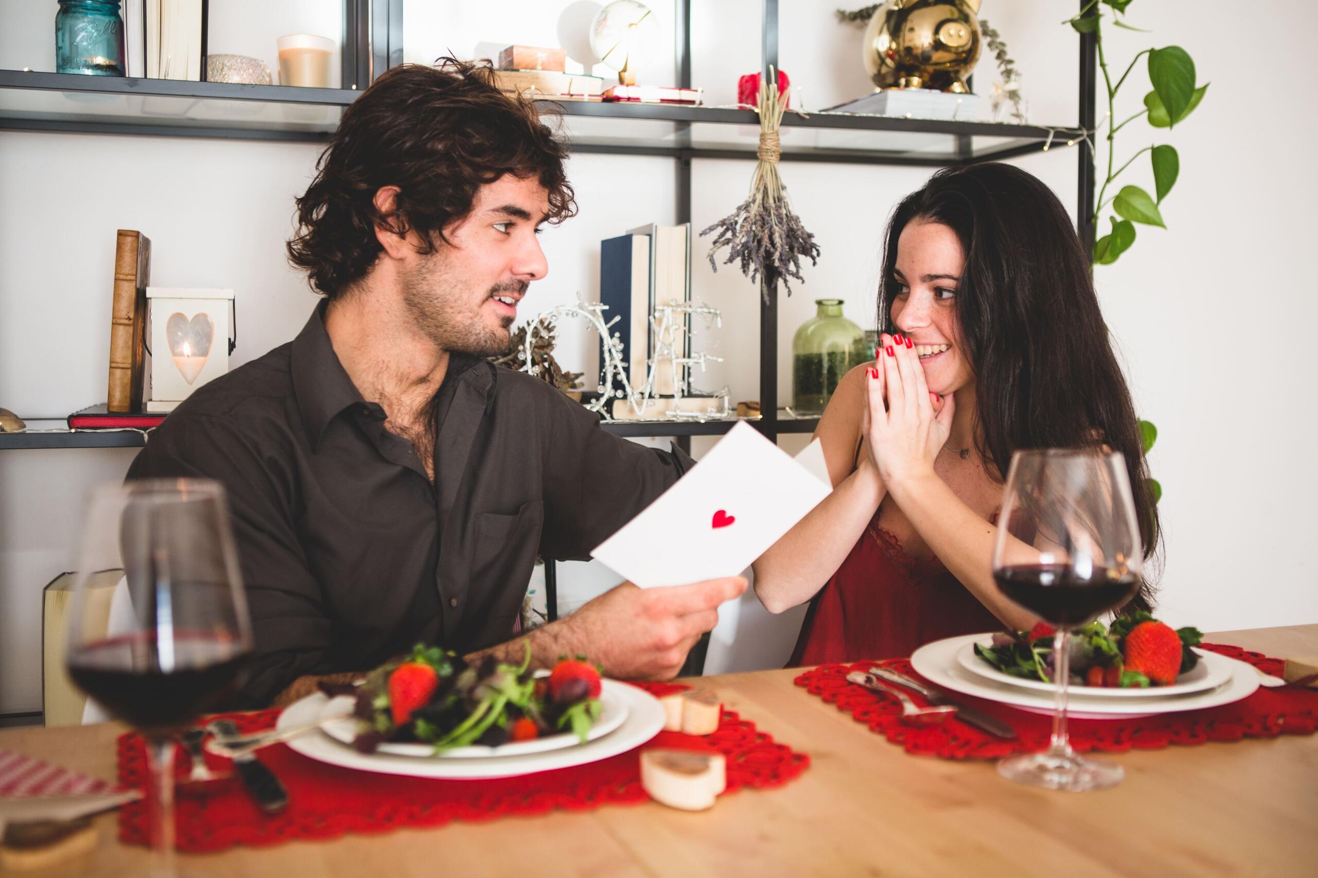 pareja con cena romantica y chico leyendole una carta en tarjeta con un corazon