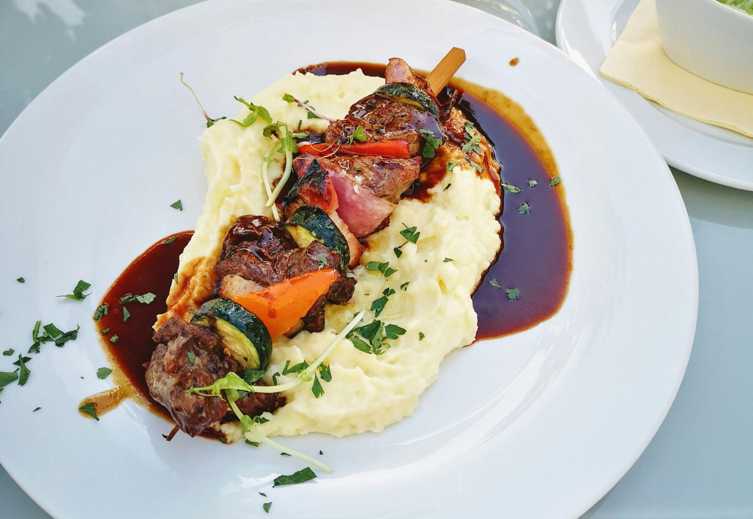 vista de un plato de pure con una brocheta de lomo al juego y vegetales encima