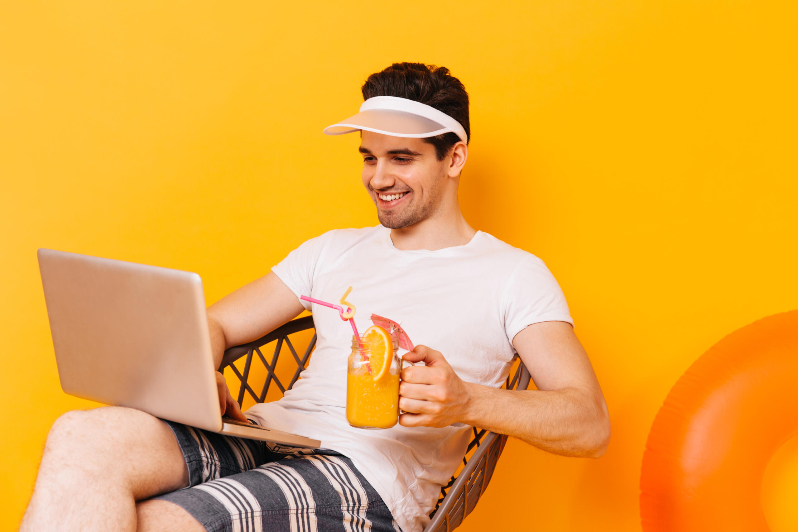 chico con look playero y un cocktail en la mano sentado sonriente mirando su laptop