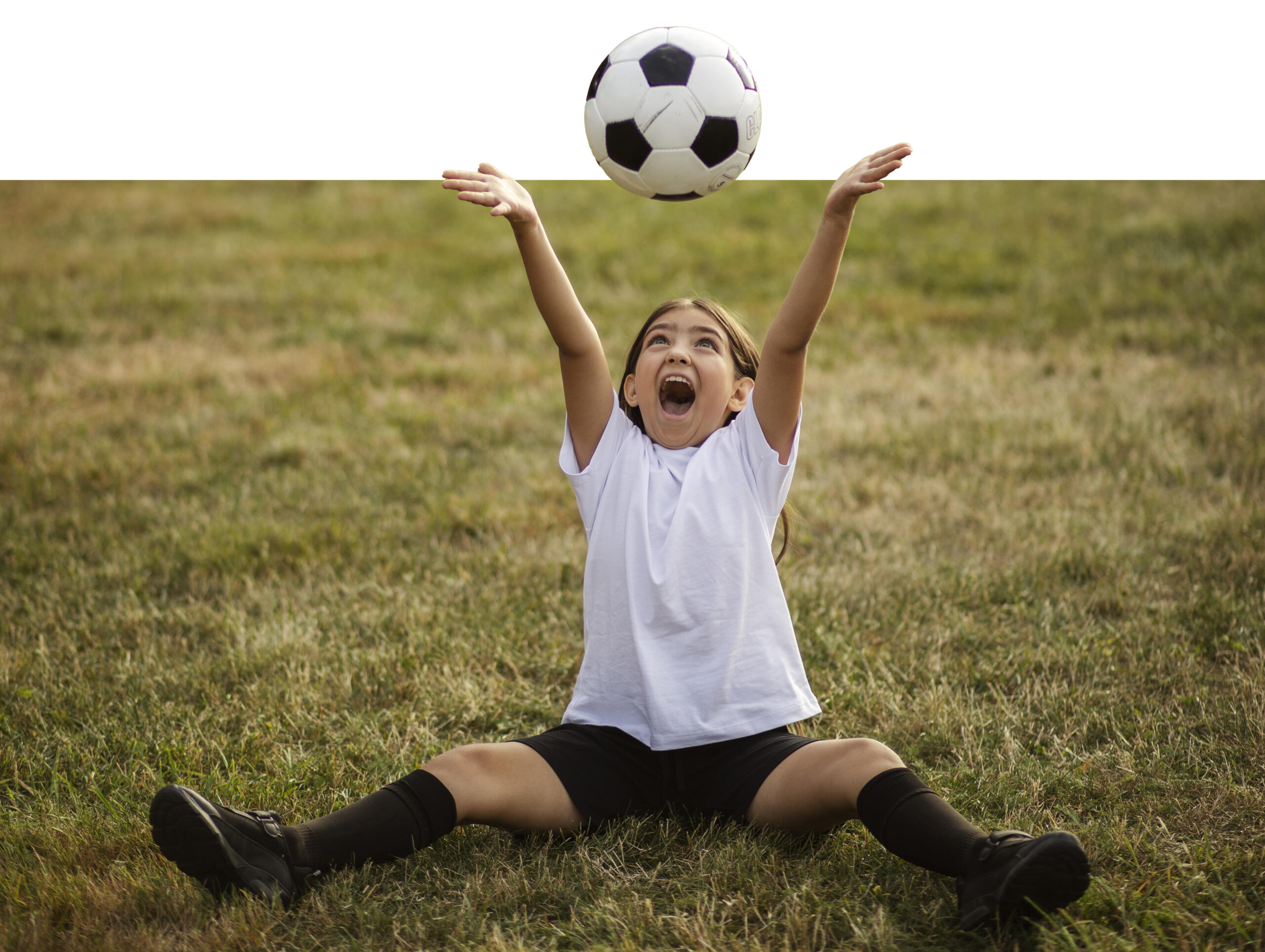 niña sentada en la cancha sonriente y con los brazos extendido hacia arriba para atrapar la pelota de futbol