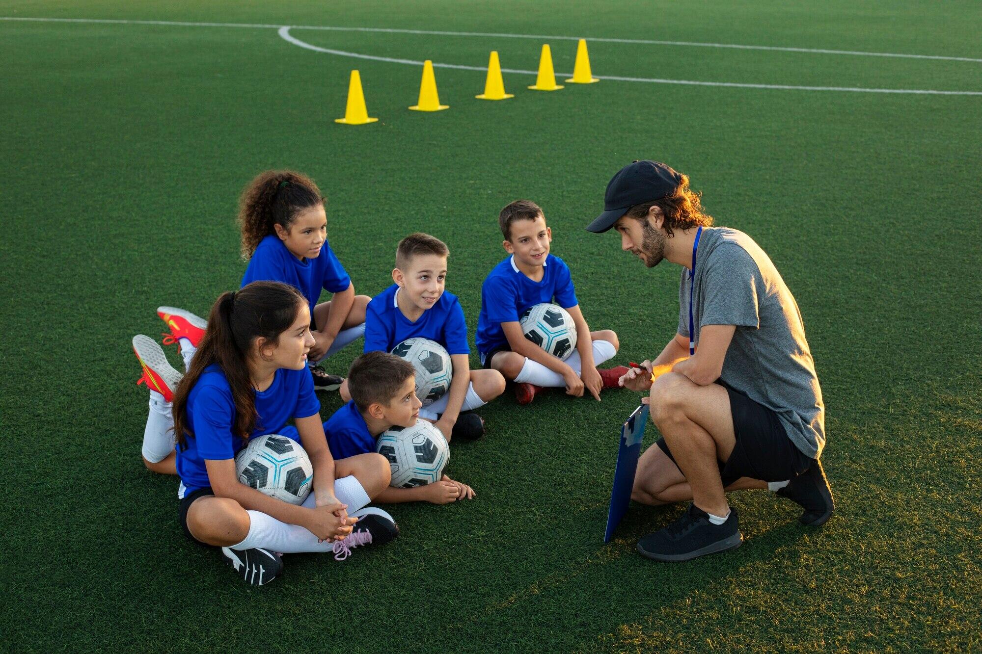 niños sentados en la cancha junto a su entrenador y con sus pelotas de futbol en mano